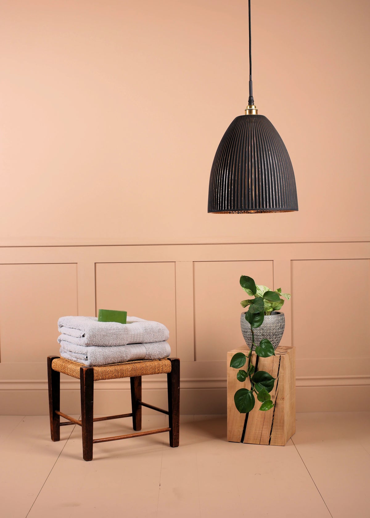 A black ribbed pendant light hangs in a styled room corner with a peach wall, next to a small stool with folded towels and a plant on a wooden block.