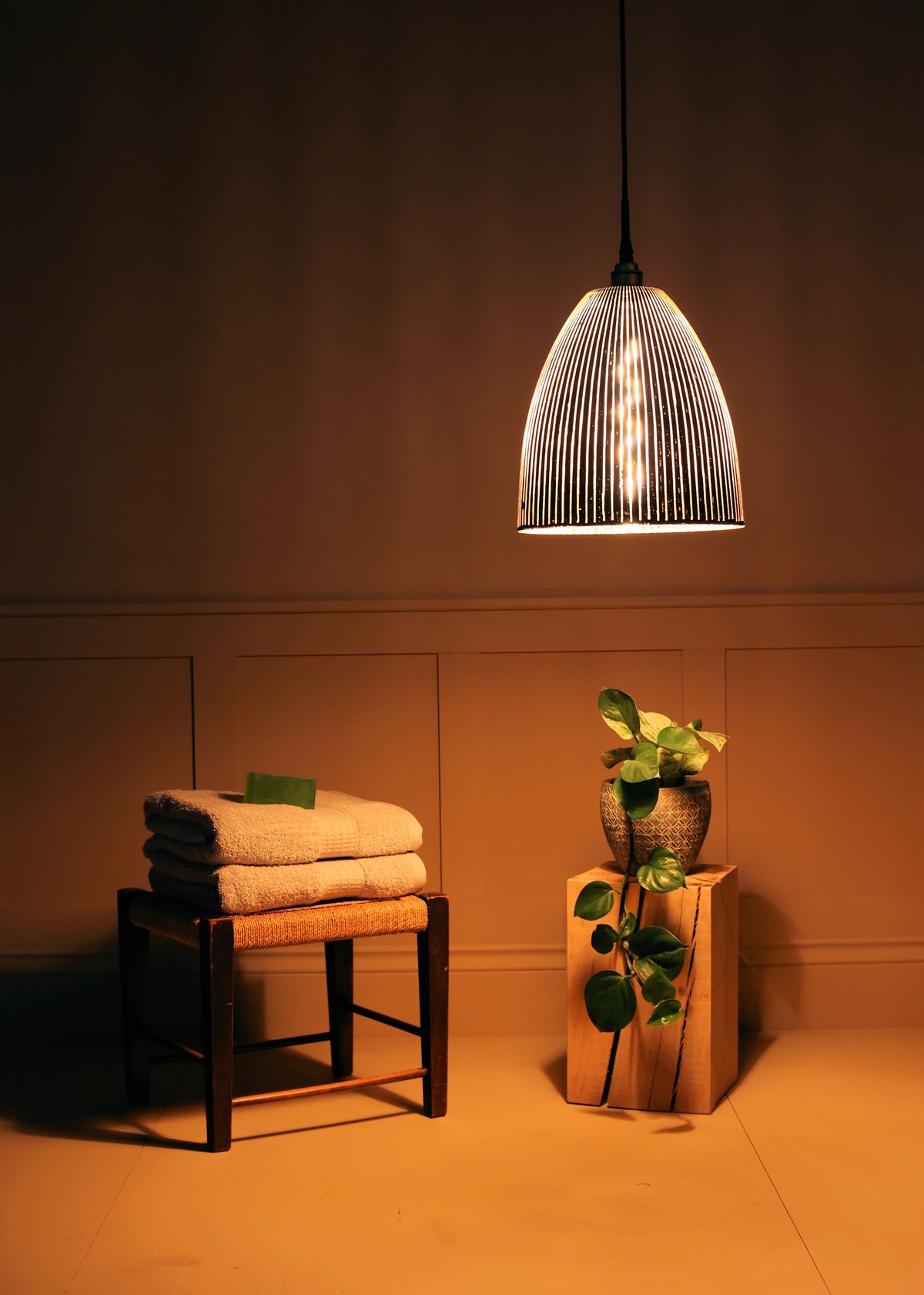 A ribbed glass pendant light glowing with warm light, hanging in a spa-like room setting with folded towels on a stool and a potted plant on a wooden block.