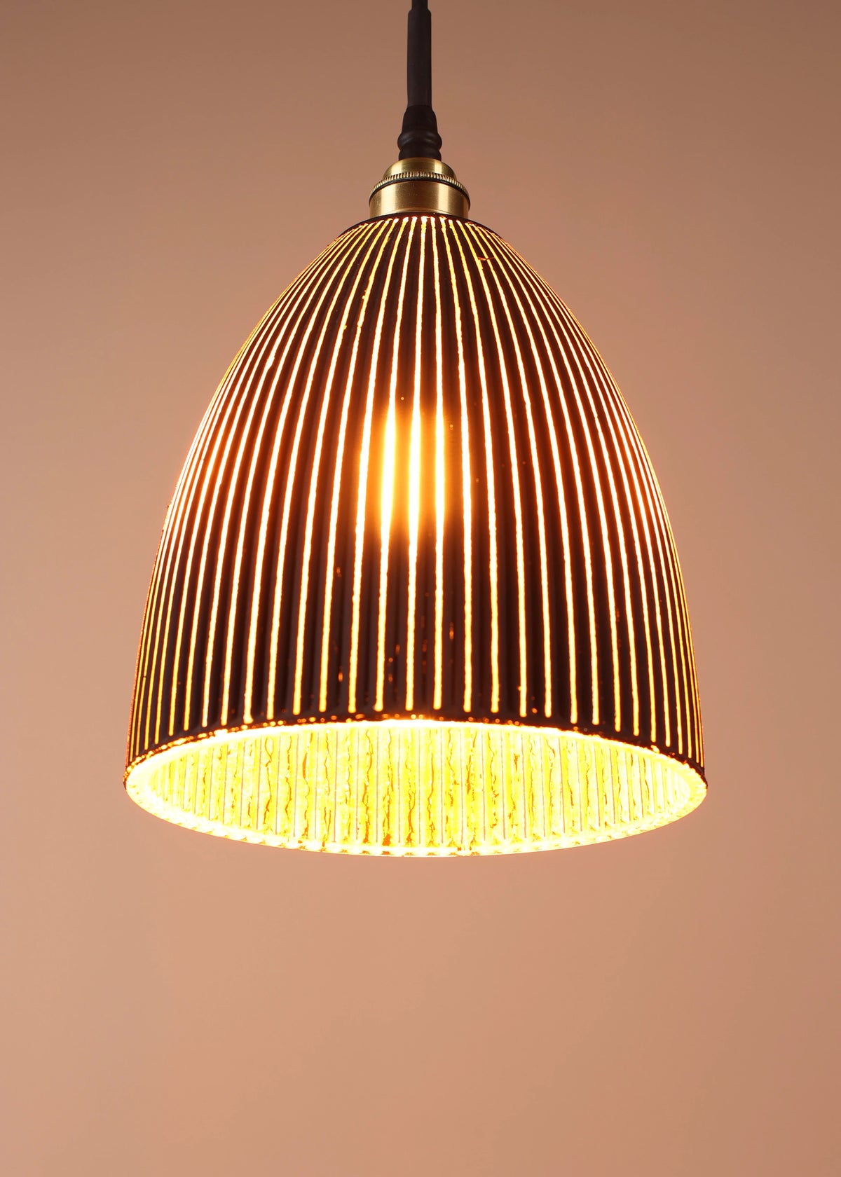 A dome-shaped pendant light with a warm brass or bronze-toned metal frame and vertical slats, glowing with a warm yellow light against a plain beige background.