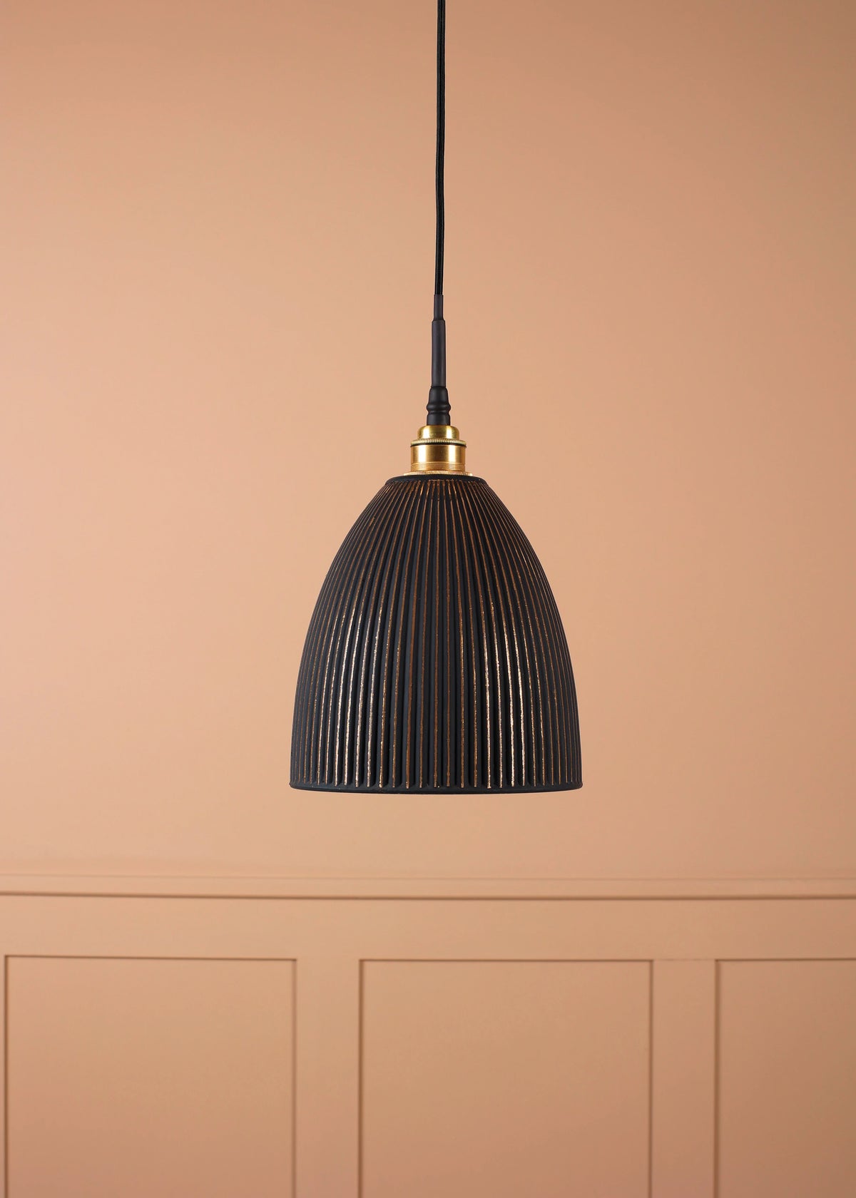 A black ribbed dome pendant light hanging from a black cord against a warm peach-pink paneled wall.