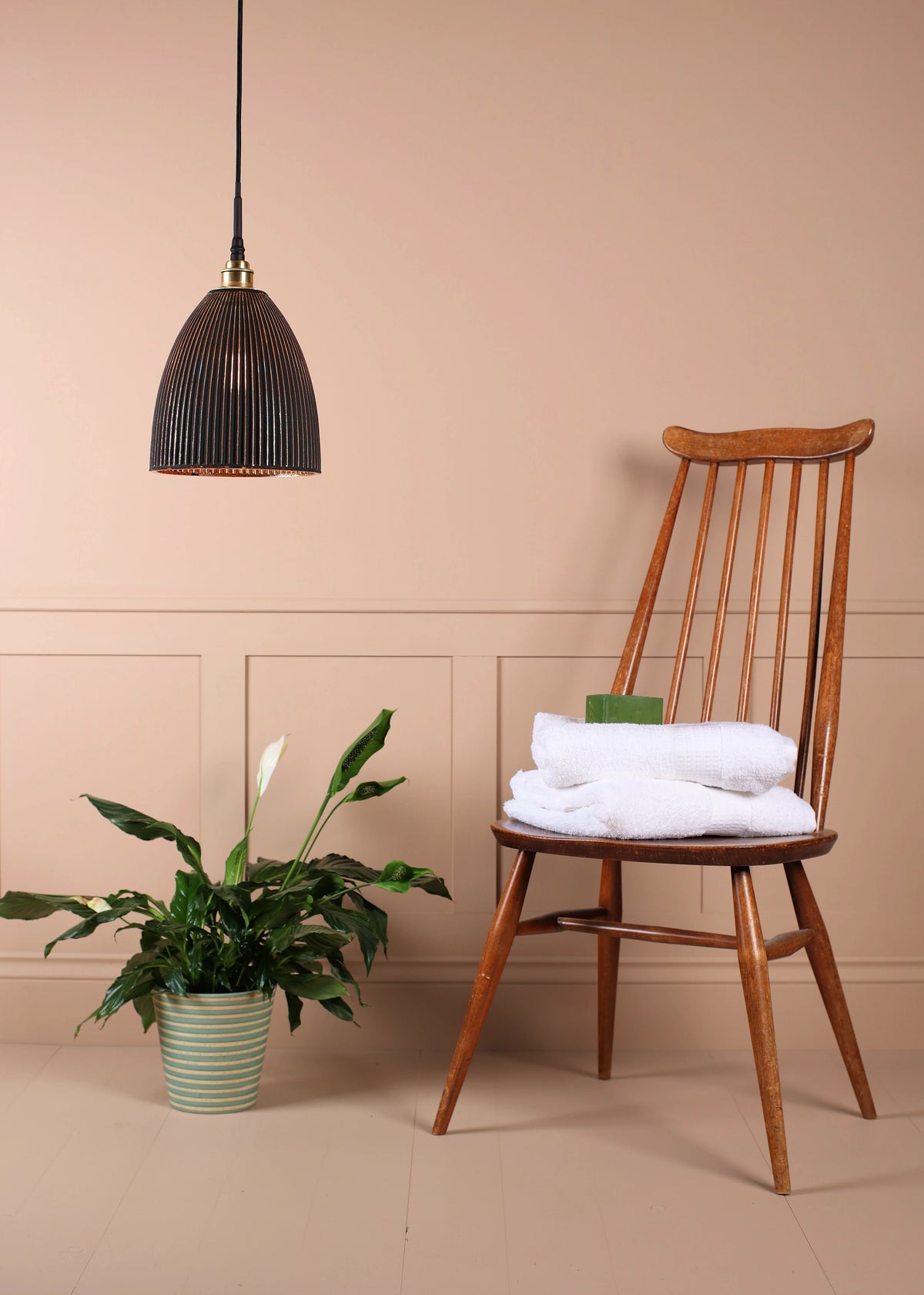 A wooden spindle-back chair with white folded towels on its seat, set against a beige wall with wainscoting. A hanging dark ribbed pendant light is on the left, and a potted green plant sits on the floor.