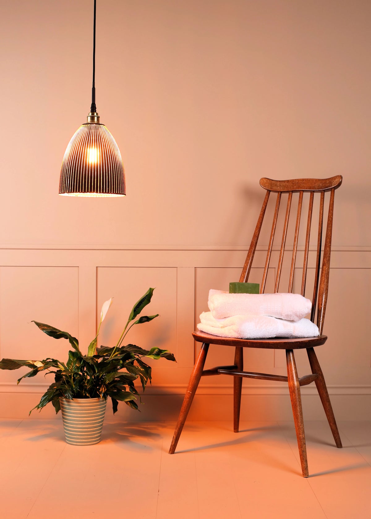 A warm, cozy interior scene featuring a wooden spindle-back chair with folded white towels on its seat, under a hanging ribbed glass pendant light. A potted green plant sits on the floor to the left, all set against a warm beige wall with decorative paneling.