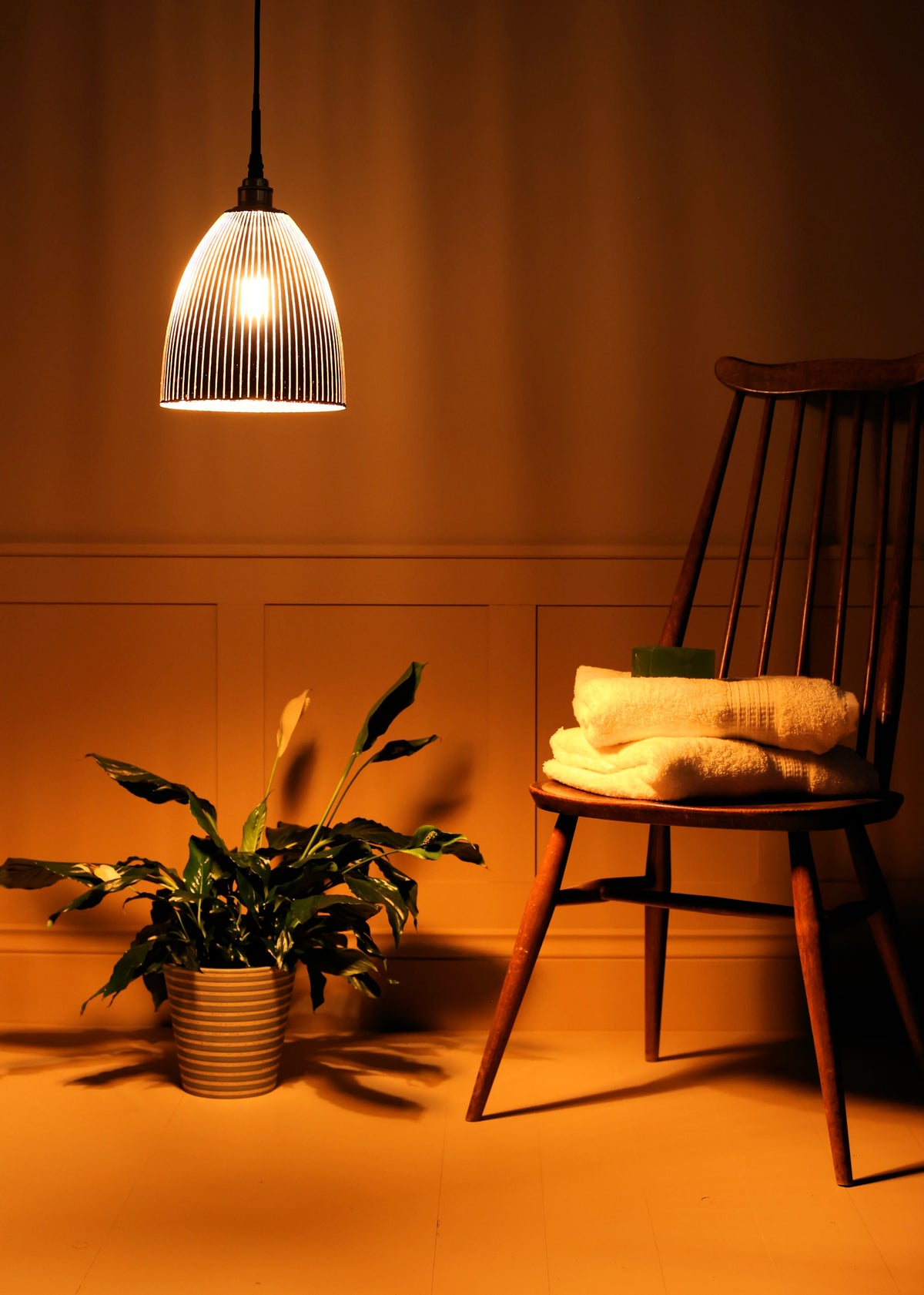 A warm, dimly lit interior scene featuring a ribbed glass pendant light glowing above a wooden chair with folded cream towels, next to a potted green plant against a dark wood-paneled wall.