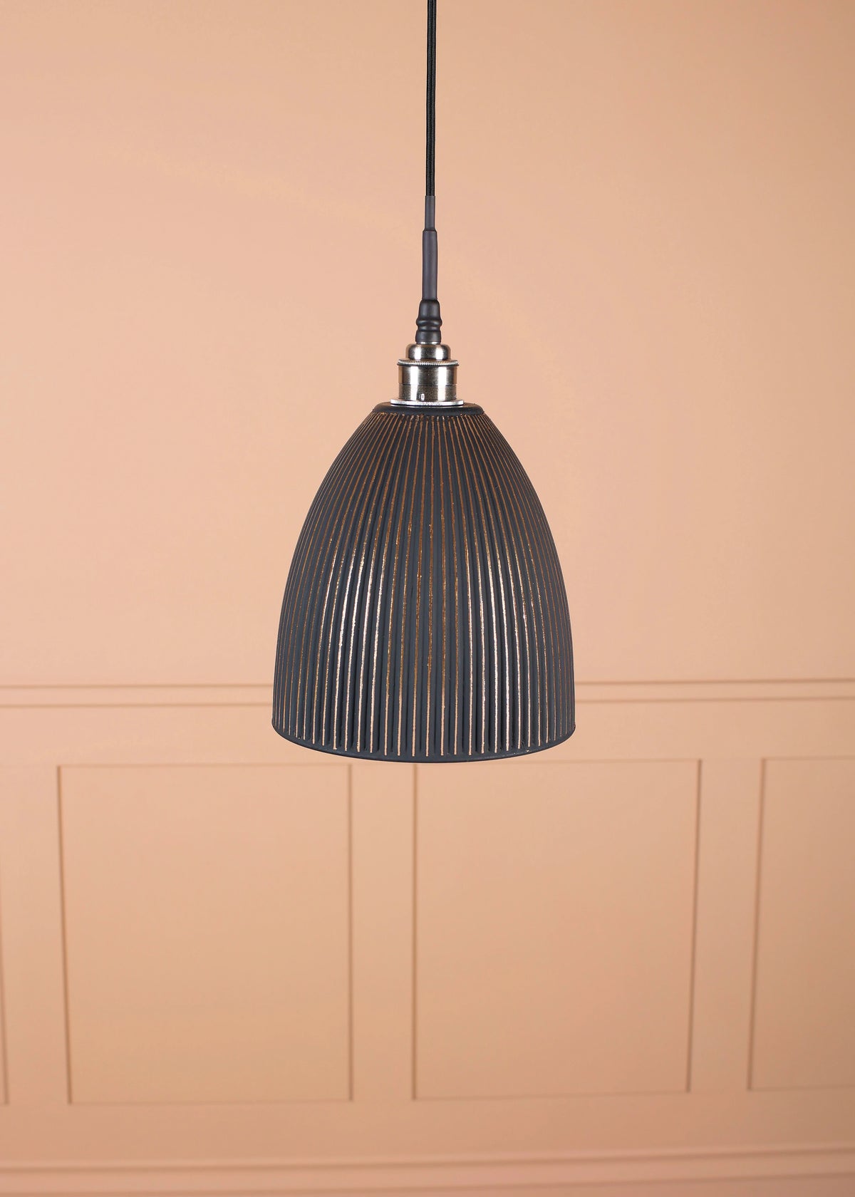 A black ribbed pendant light hanging from a black cord against a warm peach-colored paneled wall.