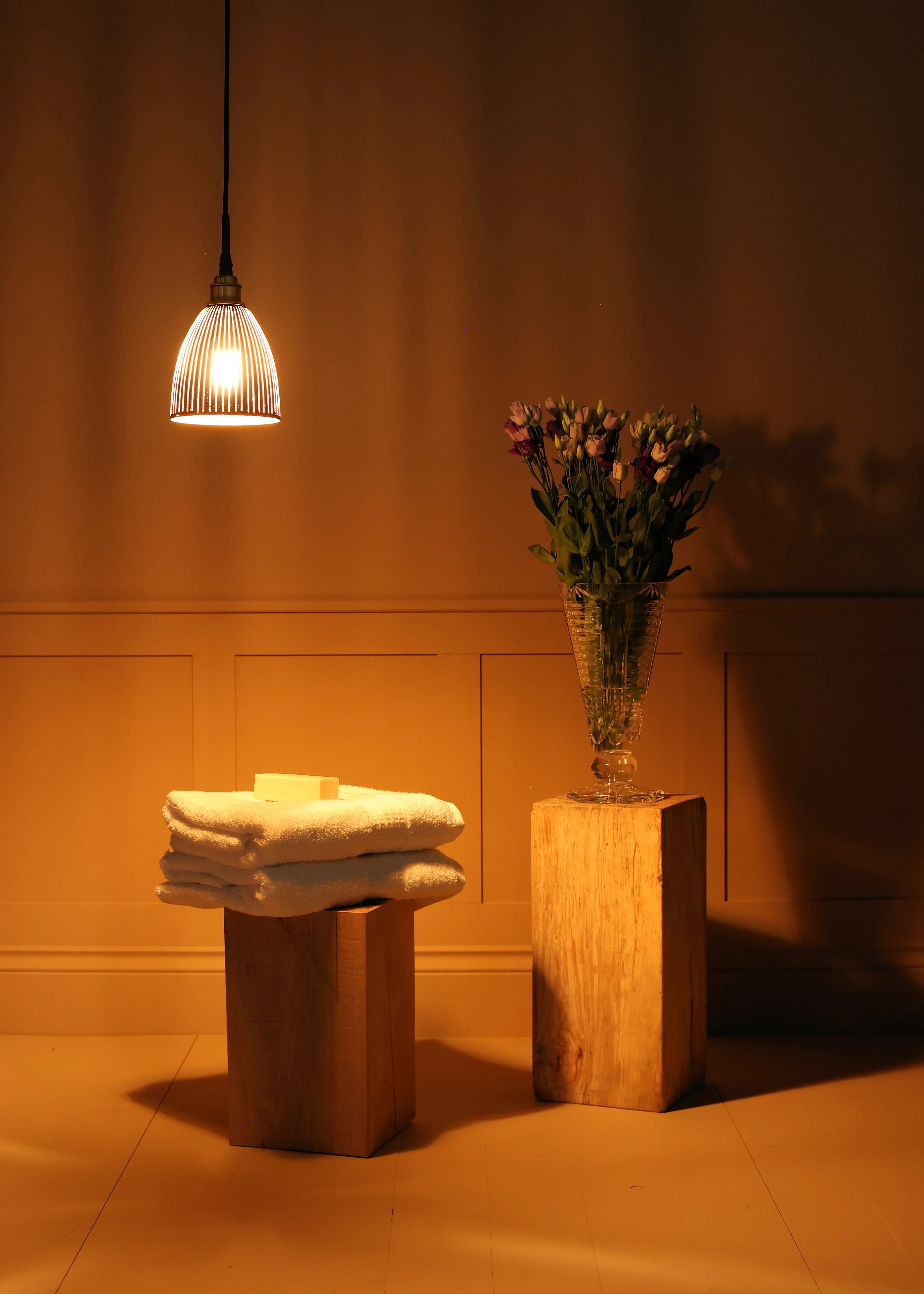 Black & gold bell shaped bathroom pendant light, stack of white towels on a wooden stool with a vase of flowers in dimly lit room