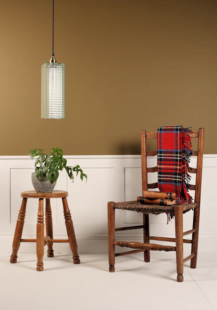 Wooden chair with a plaid blanket, wooden stool with plant, and pendant light against a beige wall.