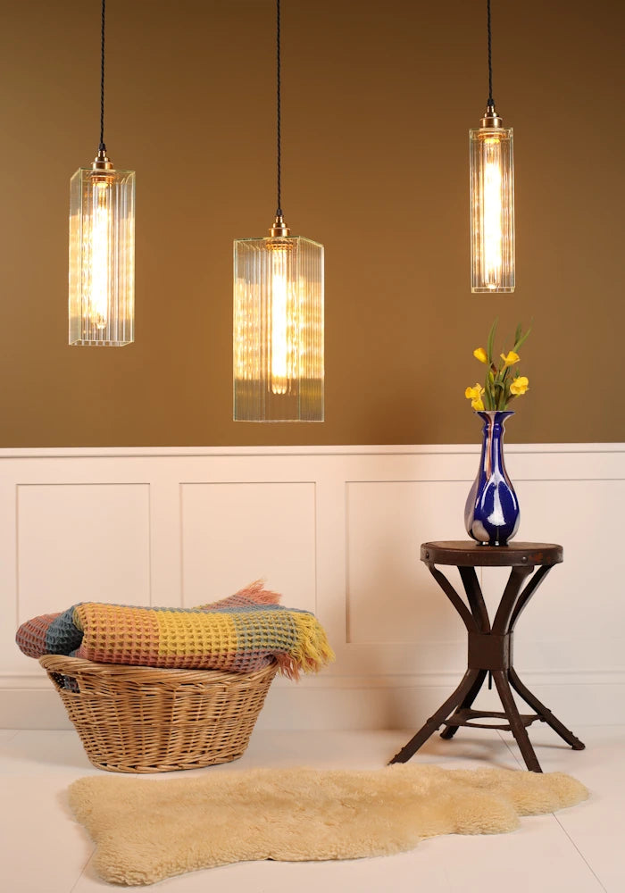 Three pendant lights hanging above a room with a wicker chair, rug, and small table with a vase.
