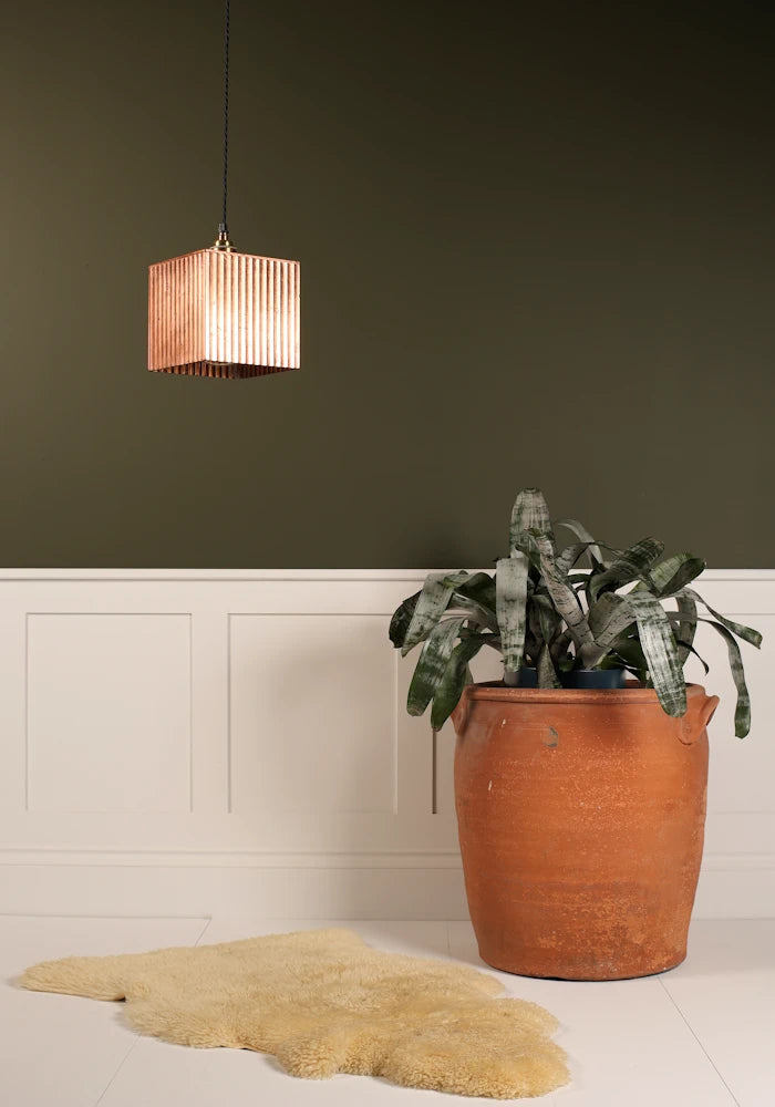 Square copper pendant light hanging over imitation fur rug, large terracotta plant pot on a green and white background.