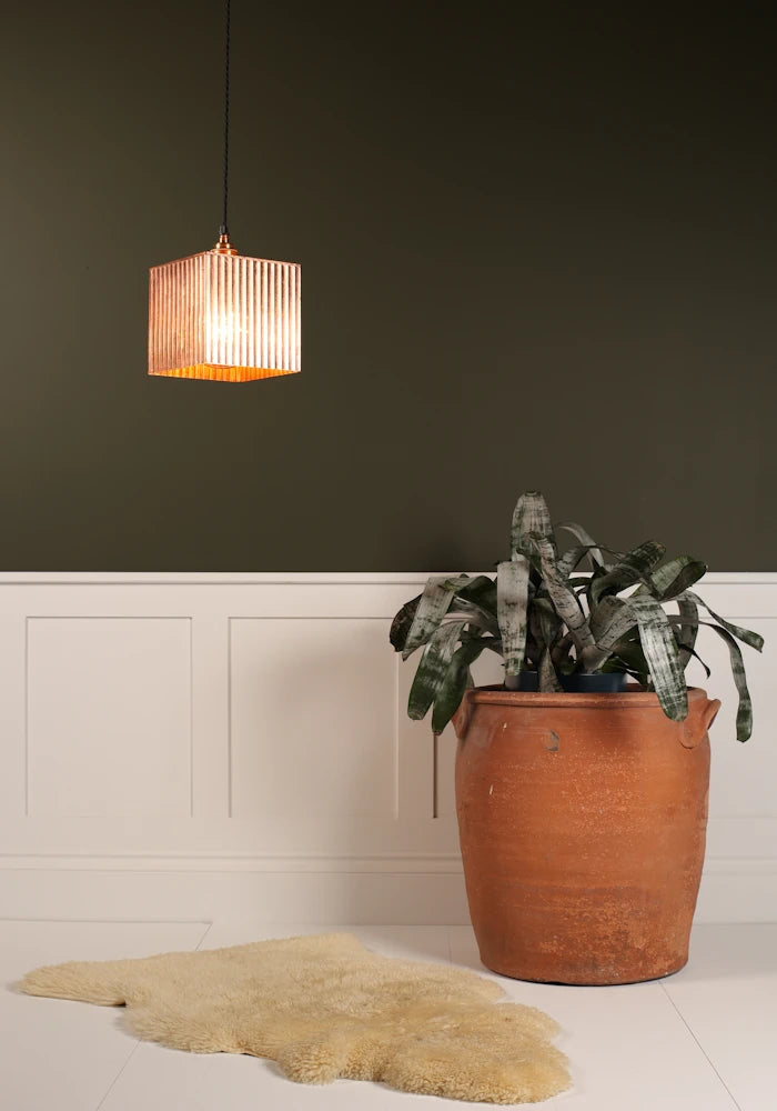 Square copper pendant light hanging over imitation fur rug, large terracotta plant pot on a green and white background.