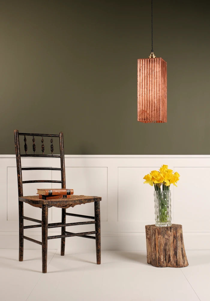 Large statement copper leaf pendant light hanging over vase of daffodils, wooden chair with white and sage green background.