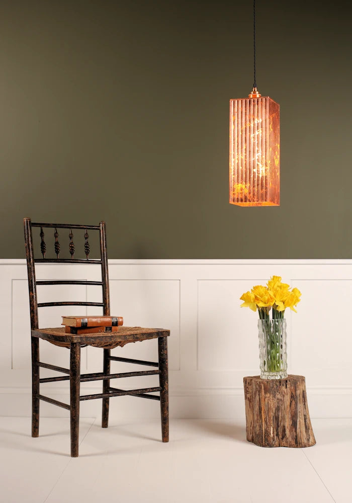 Large statement copper leaf pendant light hanging over vase of daffodils, wooden chair with white and sage green background.