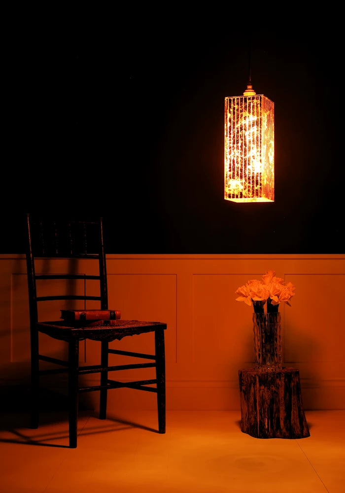 Large statement copper leaf pendant light hanging over vase of daffodils, wooden chair in dimly lit room.