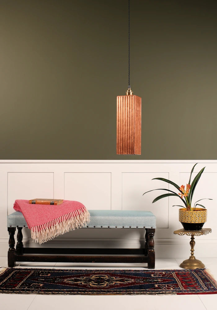 Feature copper leaf pendant light hanging over wooden bench with blanket and plant. White and sage green background.
