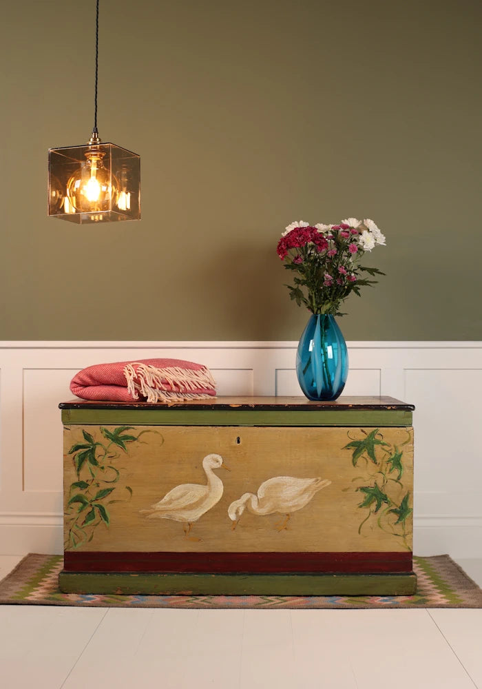 Decorative pendant light, ornate chest with swan design on a wooden floor against a green wall.