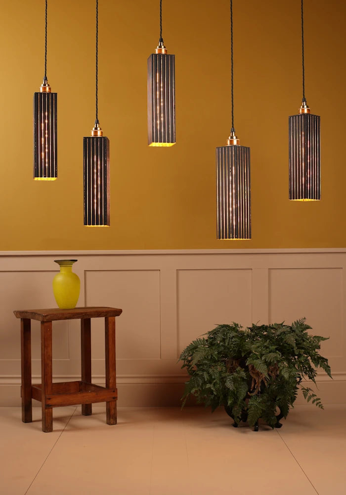 Five pendant lights hanging against a mustard yellow wall with a wooden side table and plant in the foreground.