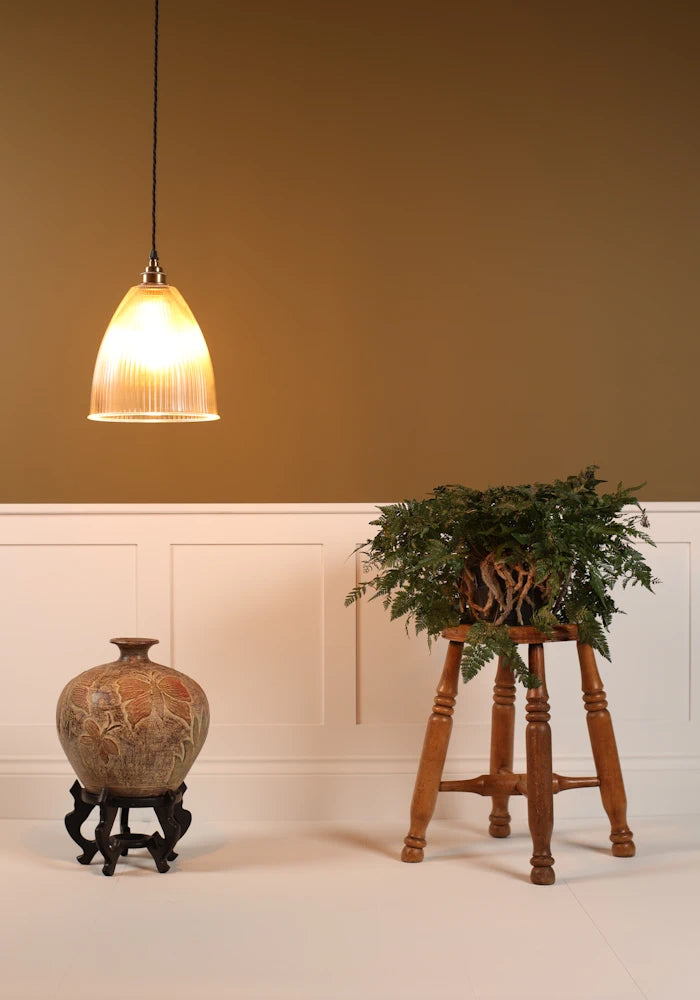Bell shaped prismatic glass pendant light hanging over decorative vase, stool with green plant, beige background