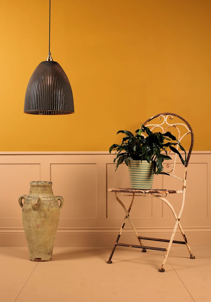 Room interior with a black pendant light, plant on a small table, and a large vase against a yellow wall.