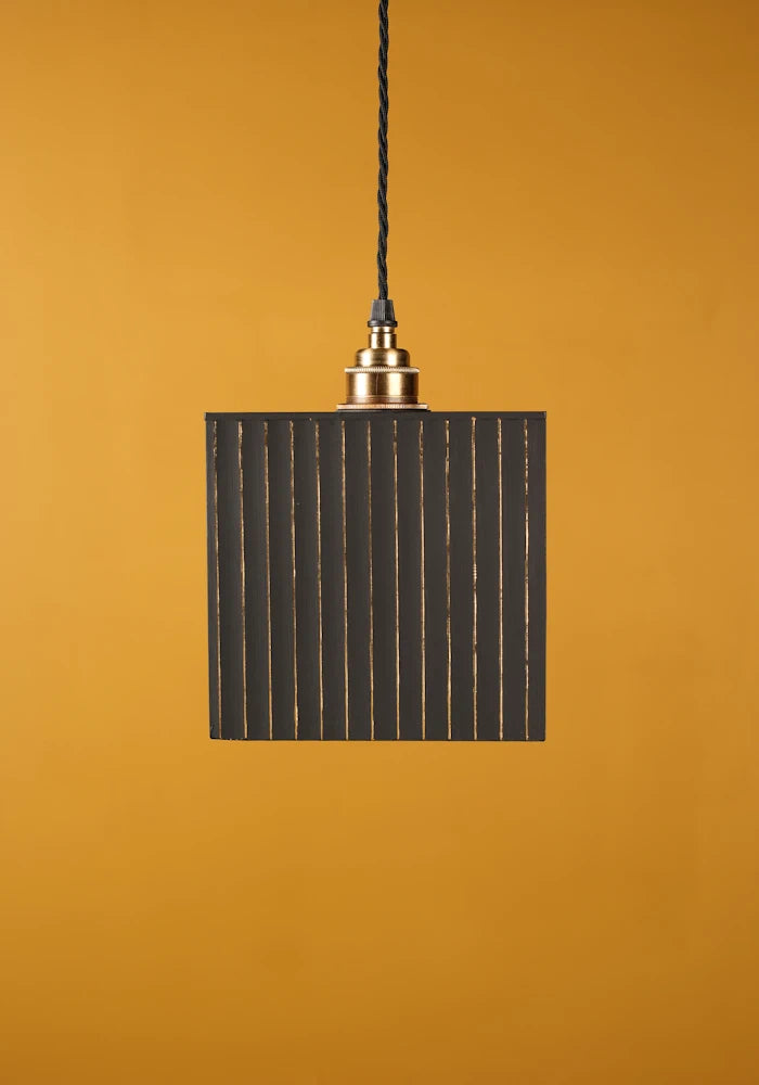 Pendant light with black rectangular shade and gold fixture against a mustard yellow background with brass lamp holder