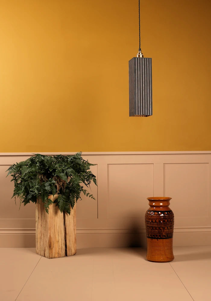 Pendant light fixture hanging above a plant and vase against a mustard yellow wall.