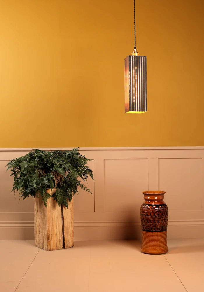 Pendant light fixture hanging above a plant and vase against a mustard yellow wall.