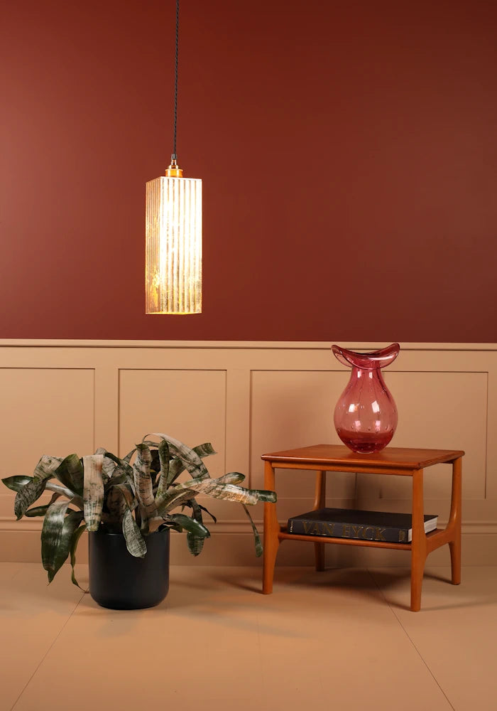 Gold pendant light fixture hanging above a plant and wooden side table with a pink vase against a red wall.