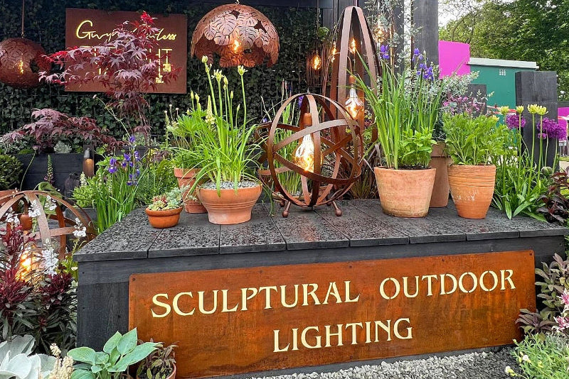 Display of sculptural outdoor lighting fixtures with plants and a sign indicating 'Sculptural Outdoor Lighting'.