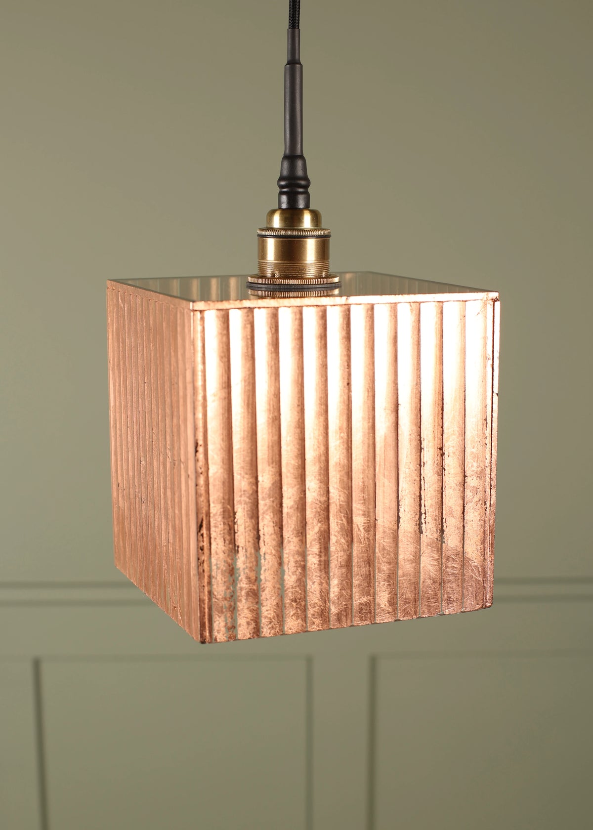 A hanging pendant light with a square, vertically fluted metallic shade in a warm copper or rose-gold tone, suspended from a black cord with a brass-colored fitting, set against a muted sage green paneled wall.