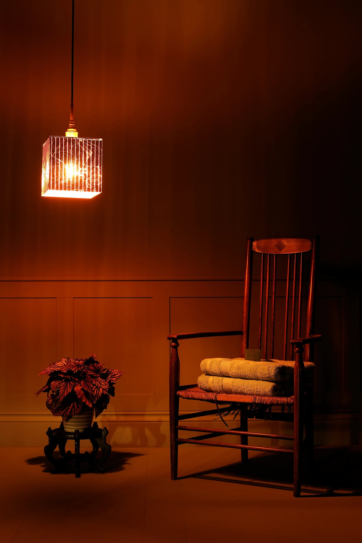 A warm, moody interior scene with a hanging cylindrical pendant light glowing orange-red, a wooden armchair with folded cream towels on the seat, and a potted plant on a small stand against paneled walls.
