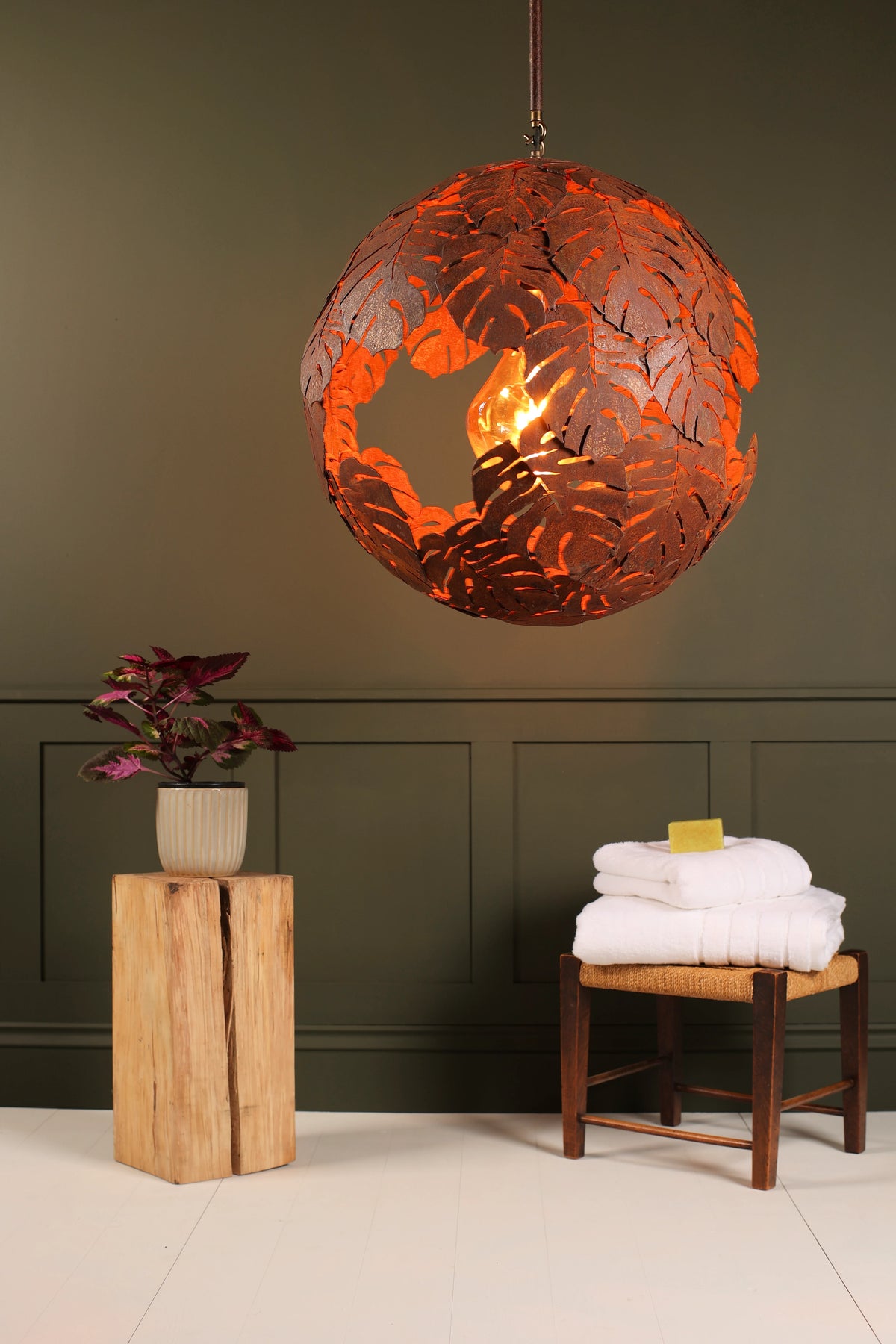 A spherical copper pendant light with a leaf cutout pattern, glowing with warm orange light, hanging in a styled bathroom or bedroom setting with a dark green paneled wall, a wooden stool with white towels, and a potted plant on a wooden block.