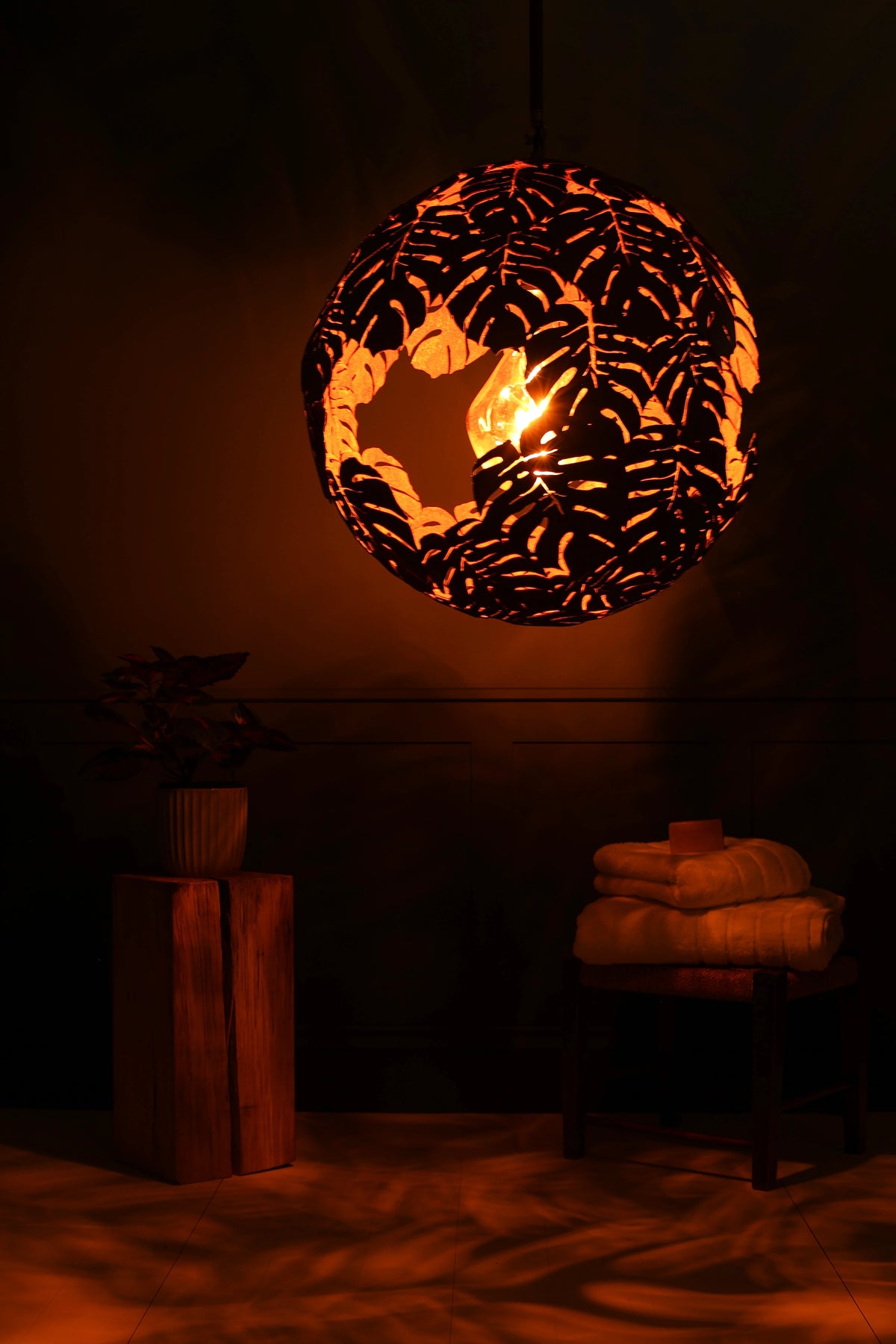 A spherical black pendant light with a leaf cutout pattern, glowing warmly from a candle inside, hanging in a dark, atmospheric room with a wooden side table and folded towels below.