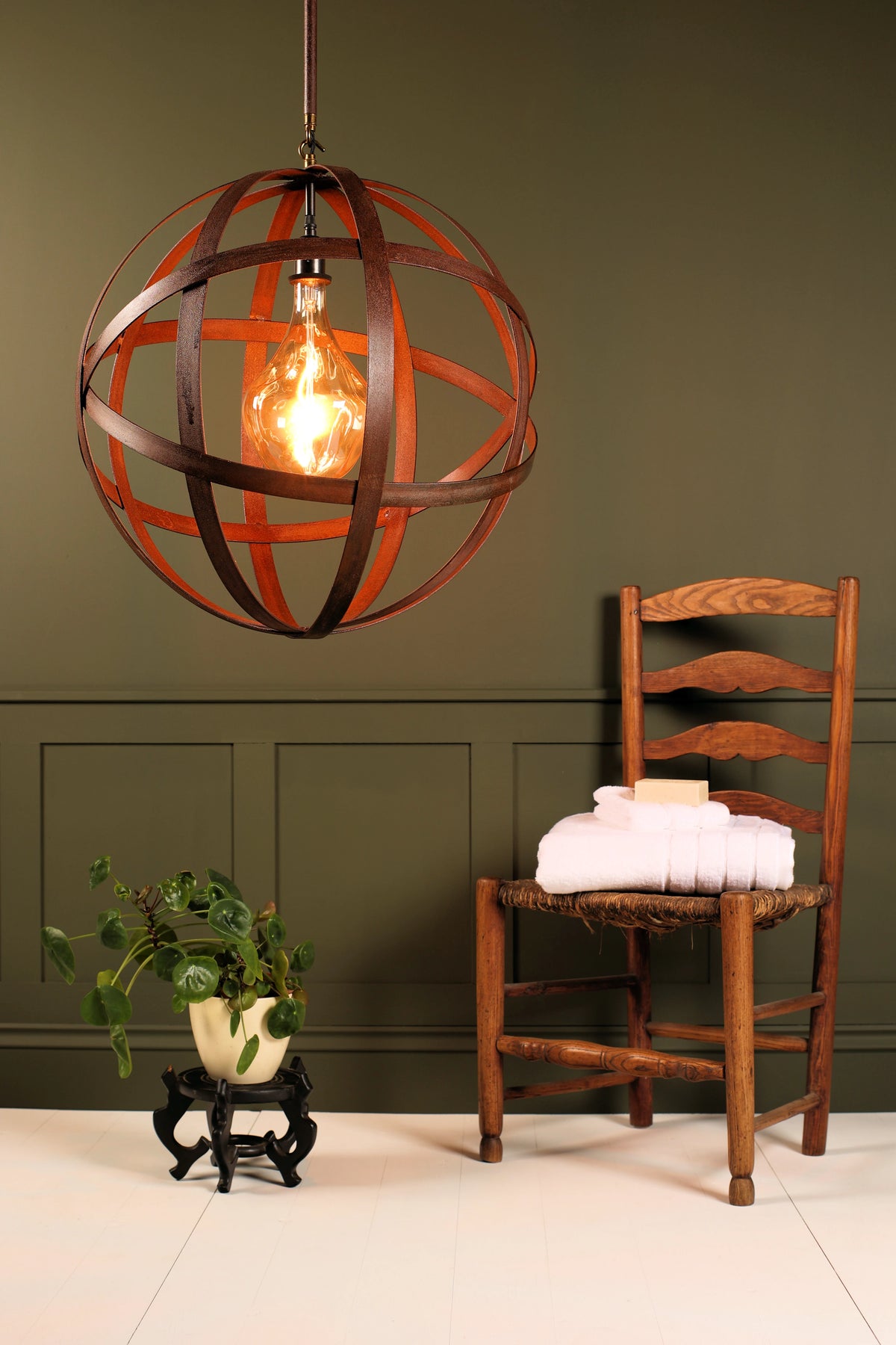 A spherical iron cage pendant light with a glowing Edison bulb hanging in a styled room setting with a wooden ladder chair and a potted plant against a dark olive green paneled wall.