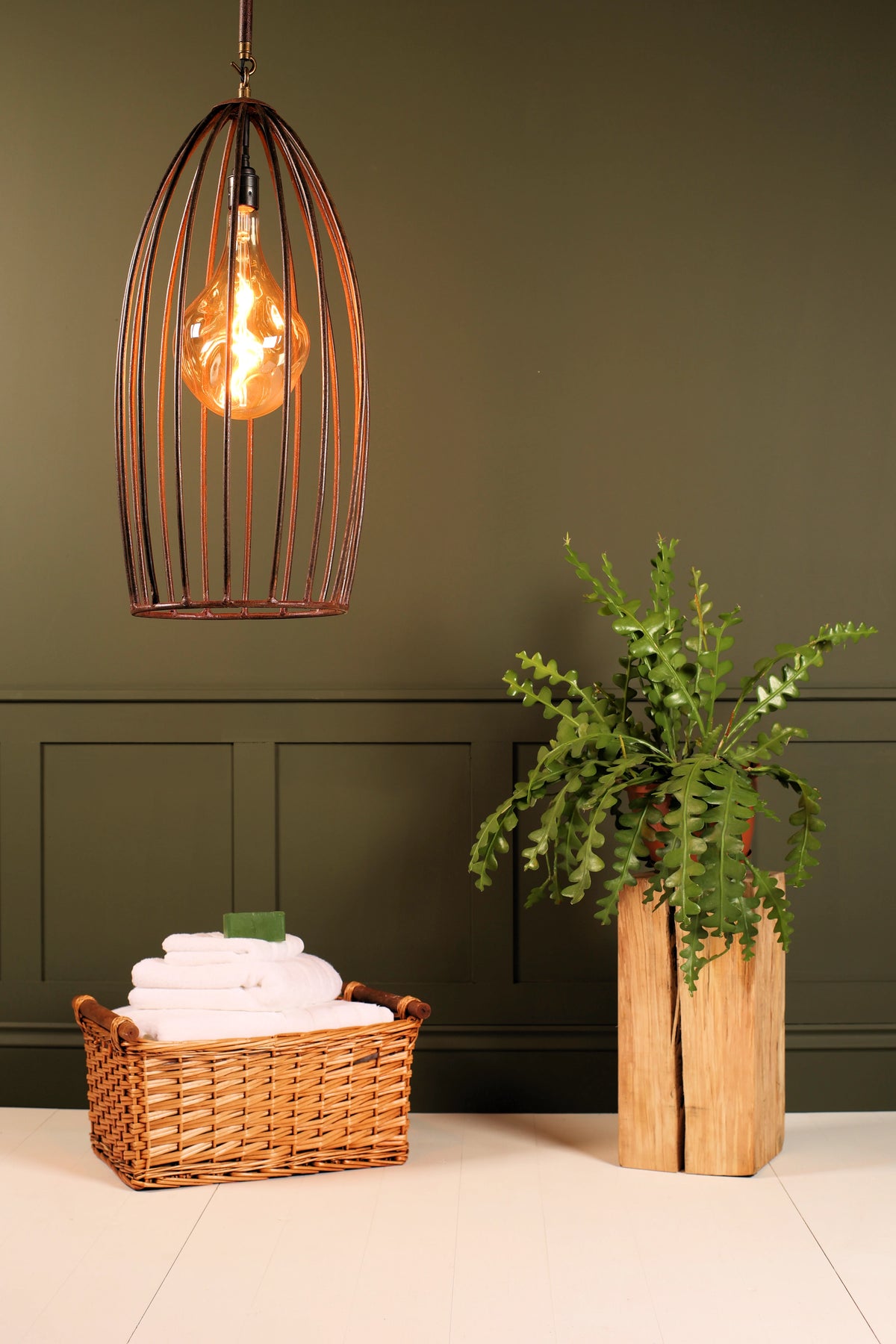 A rustic copper-toned birdcage pendant light hangs in a styled bathroom or spa setting with a dark green paneled wall, a wicker basket of folded white towels, and a green plant in a wooden vase.