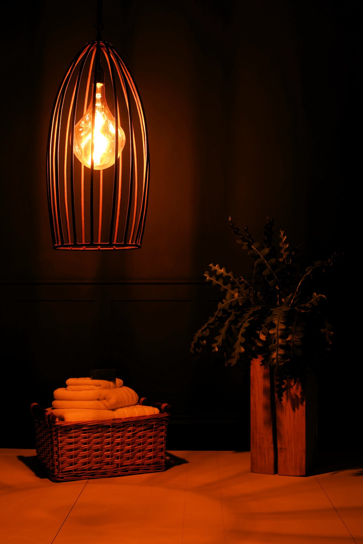 A warm, glowing birdcage pendant light hangs in a dimly lit bathroom or spa setting, casting an orange glow over folded towels in a wicker basket and a potted plant.
