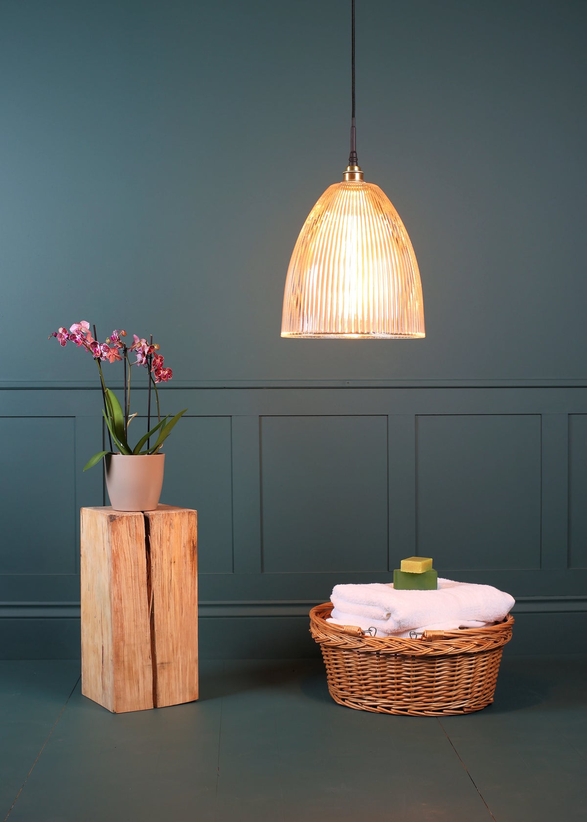 A warm, glowing pendant light with a ribbed glass shade hangs in a room. To the left, a potted orchid sits on a wooden block, and to the right, a wicker basket holds folded towels and a small green soap.