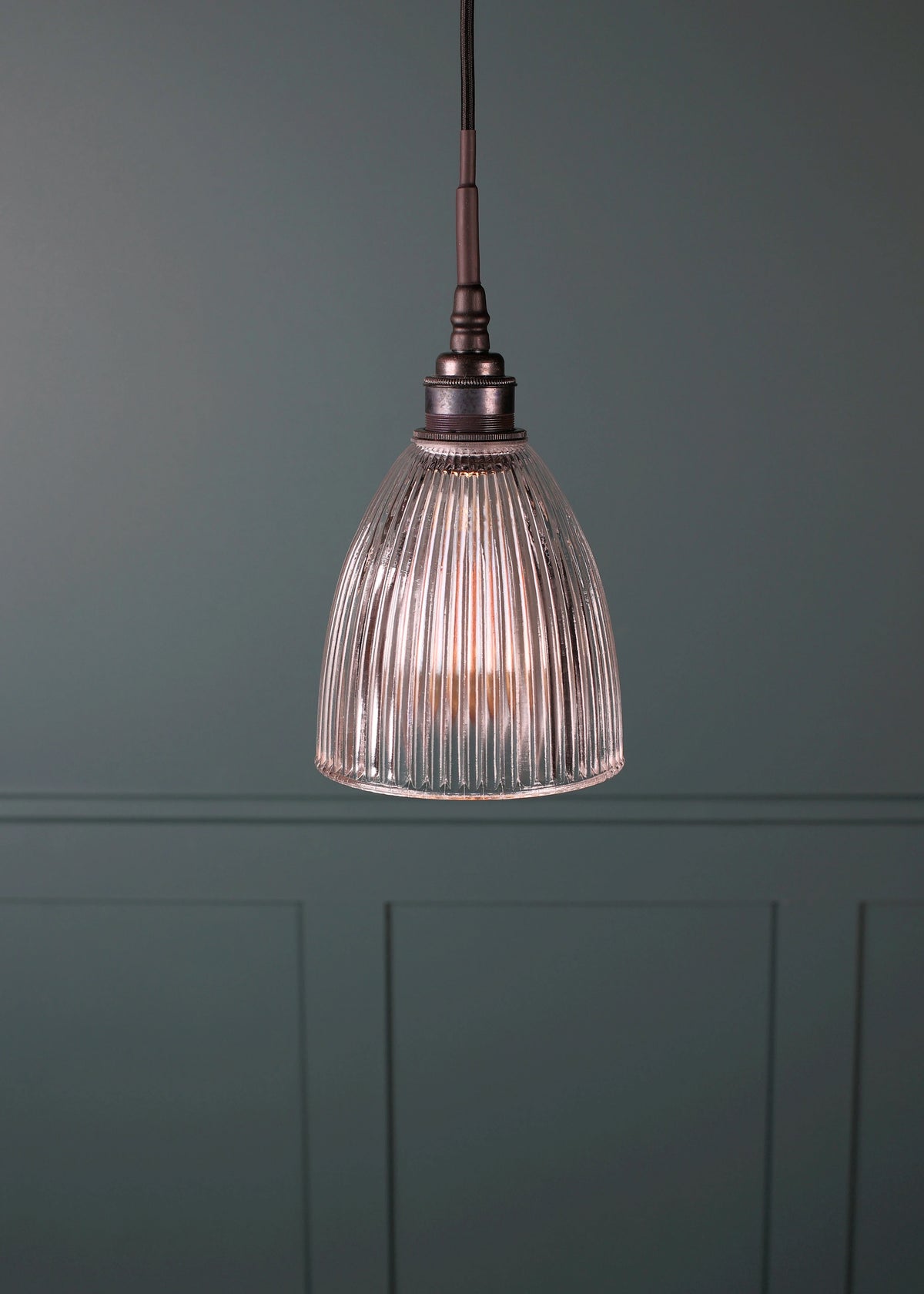 A single pendant light with a ribbed clear glass shade and a dark bronze metal top and cord, hanging against a muted green paneled wall.