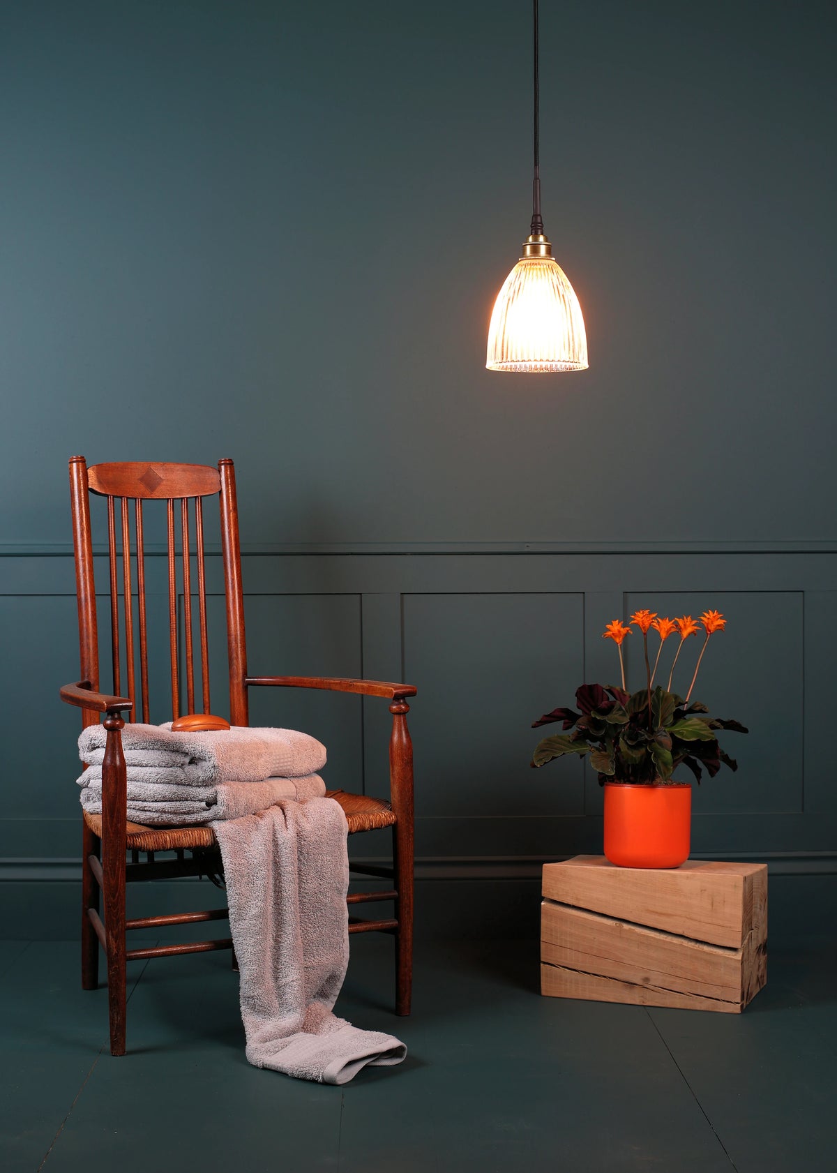 A warm-toned wooden chair in a styled room setting with a folded light gray towel on the seat and a draped throw blanket. A glowing pendant light hangs above, and a potted orange flower sits on a wooden crate against a dark teal paneled wall.