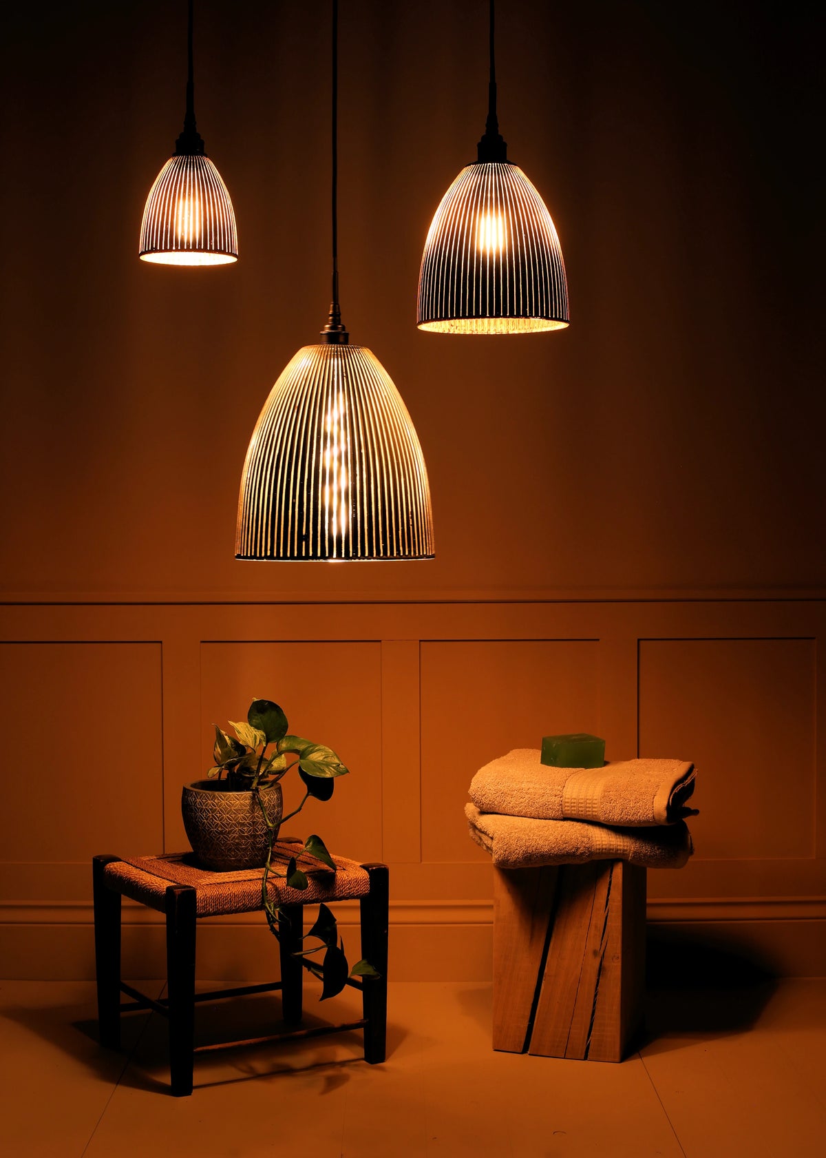 Three ribbed glass pendant light shades hanging in a warm, dimly lit bathroom or spa setting, with a small stool holding a potted plant and a folded towel on a wooden block nearby.