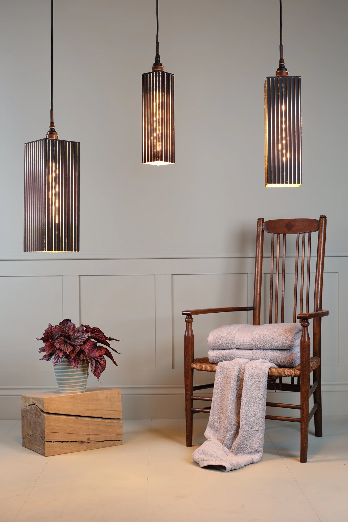 Triple black and gold bathroom pendant light cluster with plant, wooden chair and bath towels on pale green paneled background