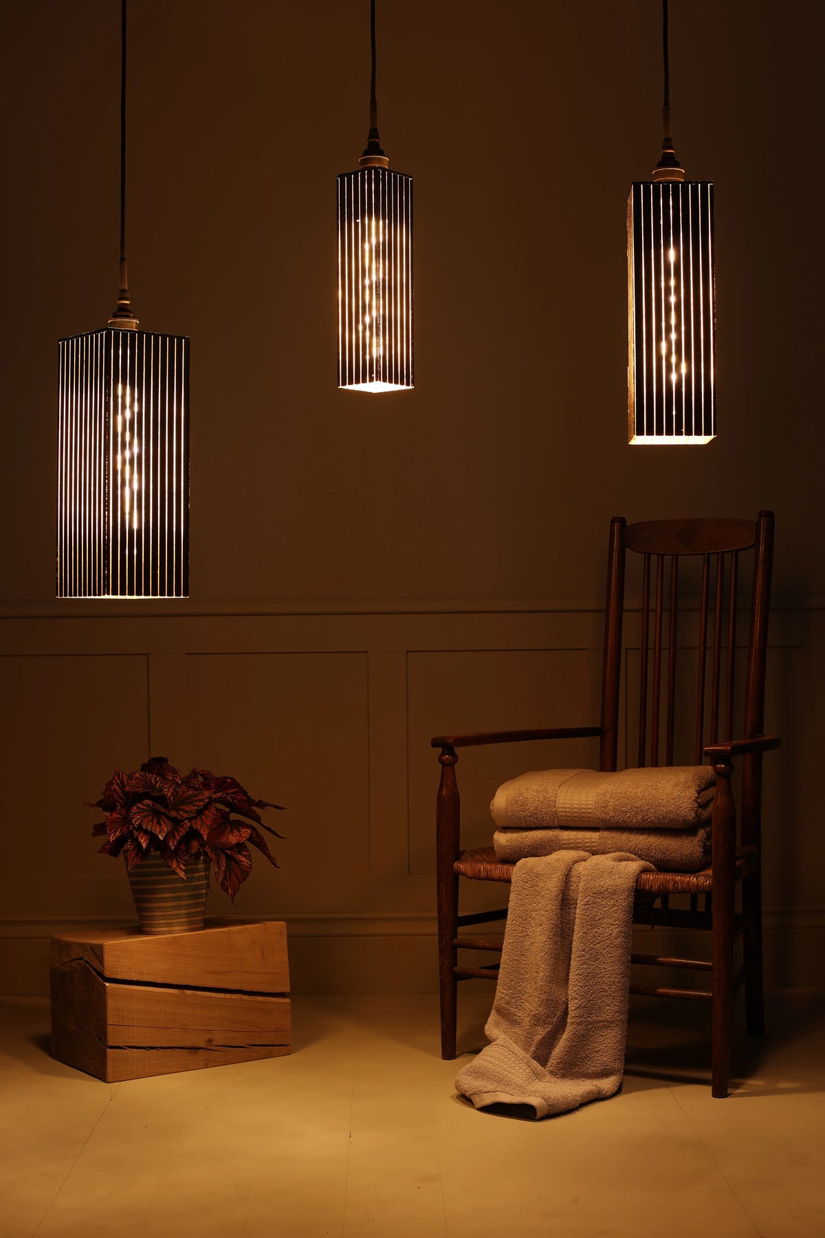 Triple black and gold bathroom pendant light cluster with plant, wooden chair and bath towels on pale green paneled background in dimly lit room