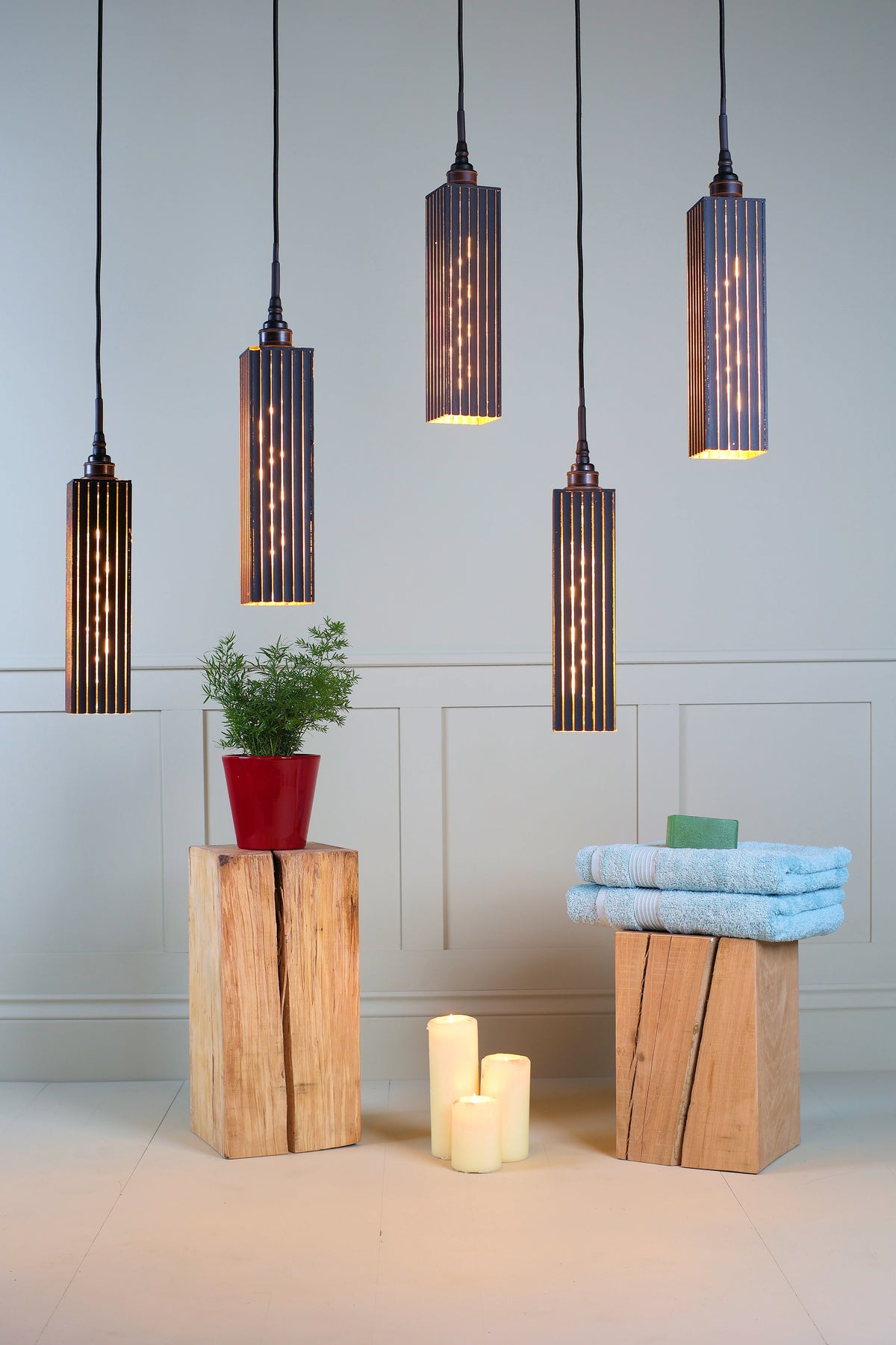 Five modern cylindrical bathroom pendant lights with dark ribbed metal shades and warm glowing bulbs, hanging in a bathroom-like setting above a light gray paneled wall. Decorative wood blocks, a potted plant, folded blue towels, and candles are arranged below.
