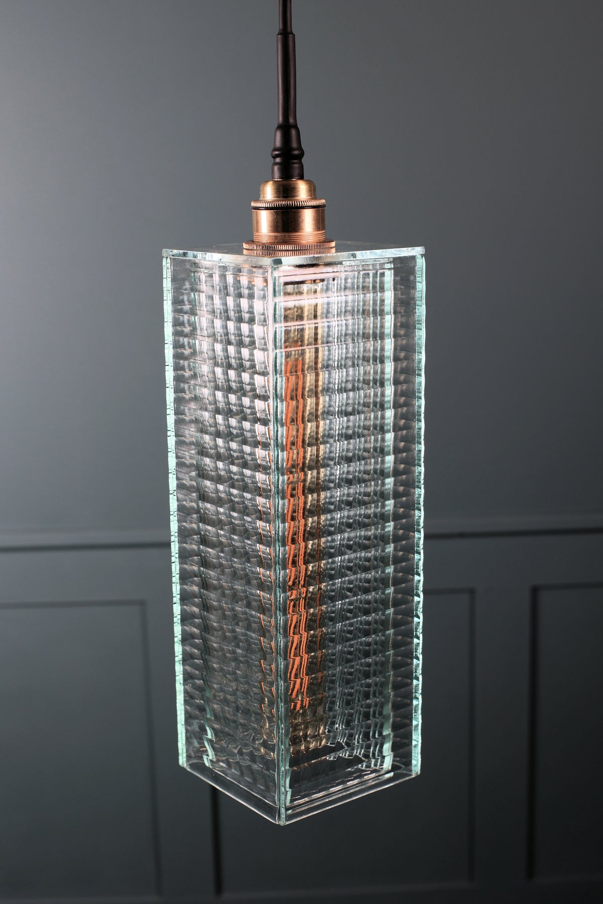 A modern pendant light with a textured, ribbed clear glass cylinder shade, a copper-finished metal cap, and a black hanging cord against a dark gray paneled wall.