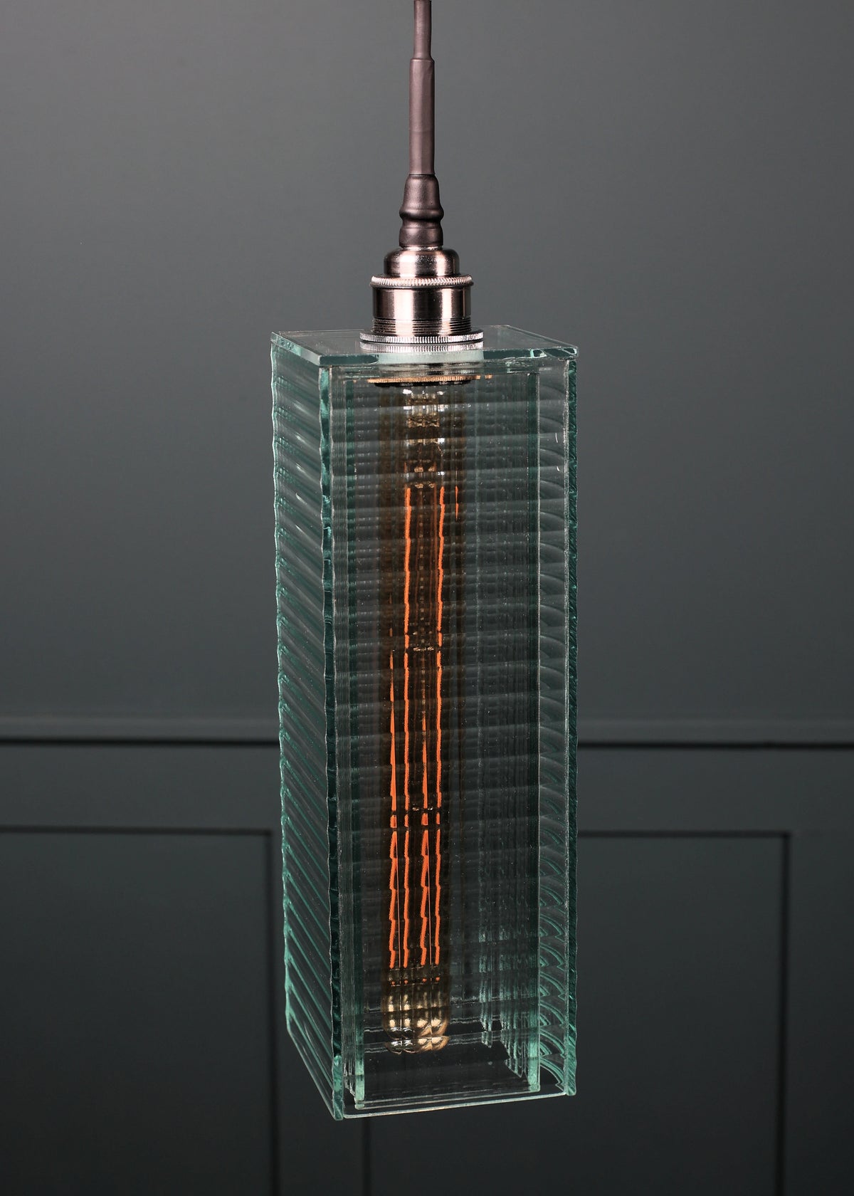 A modern rectangular pendant light with a ribbed clear glass shade, showcasing a glowing orange filament inside, suspended from a dark metal rod against a dark gray paneled background.