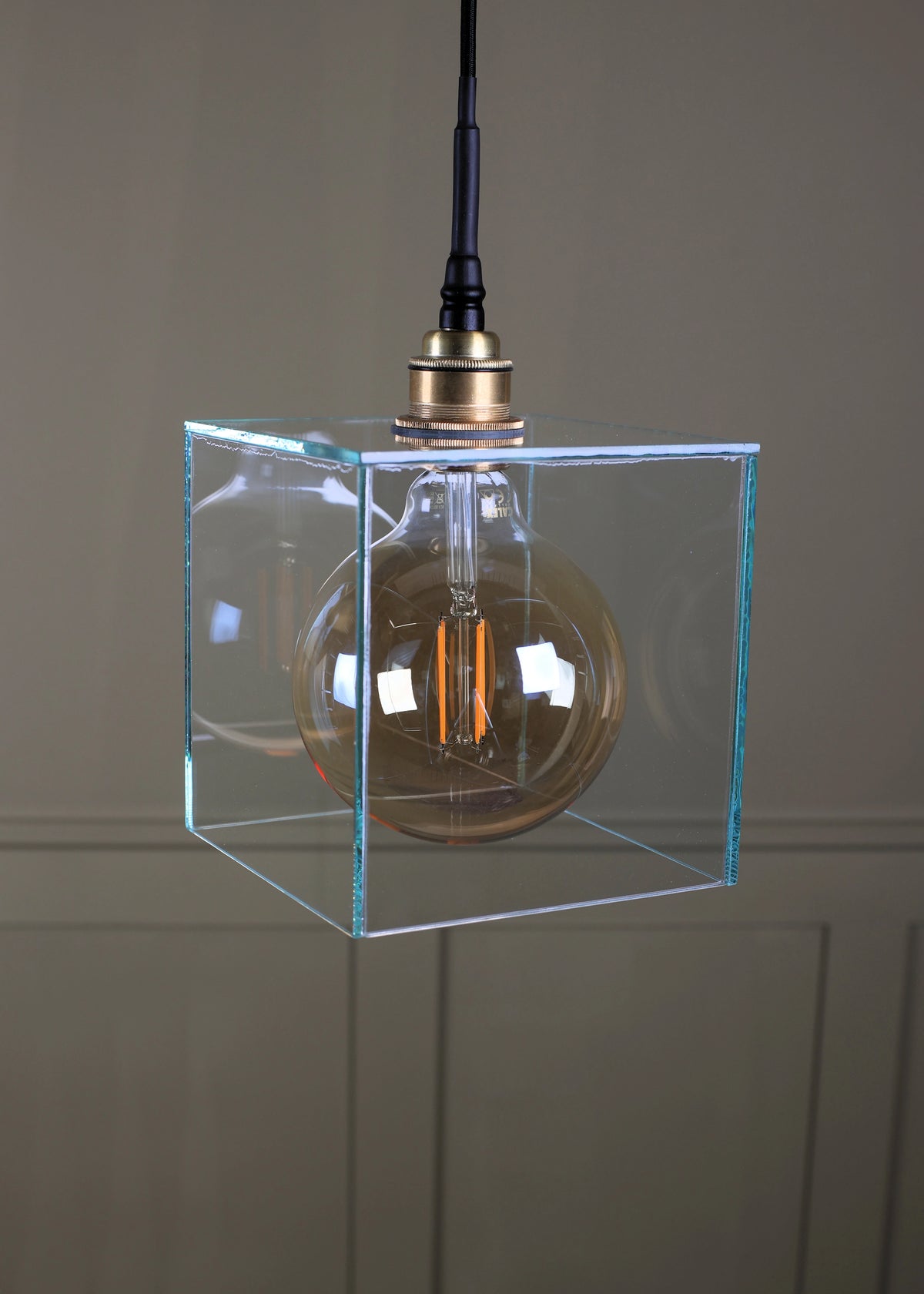 A modern pendant light featuring a clear glass cube enclosure with a warm amber glass globe bulb inside, suspended from a brass-colored metal fitting with a black cord.