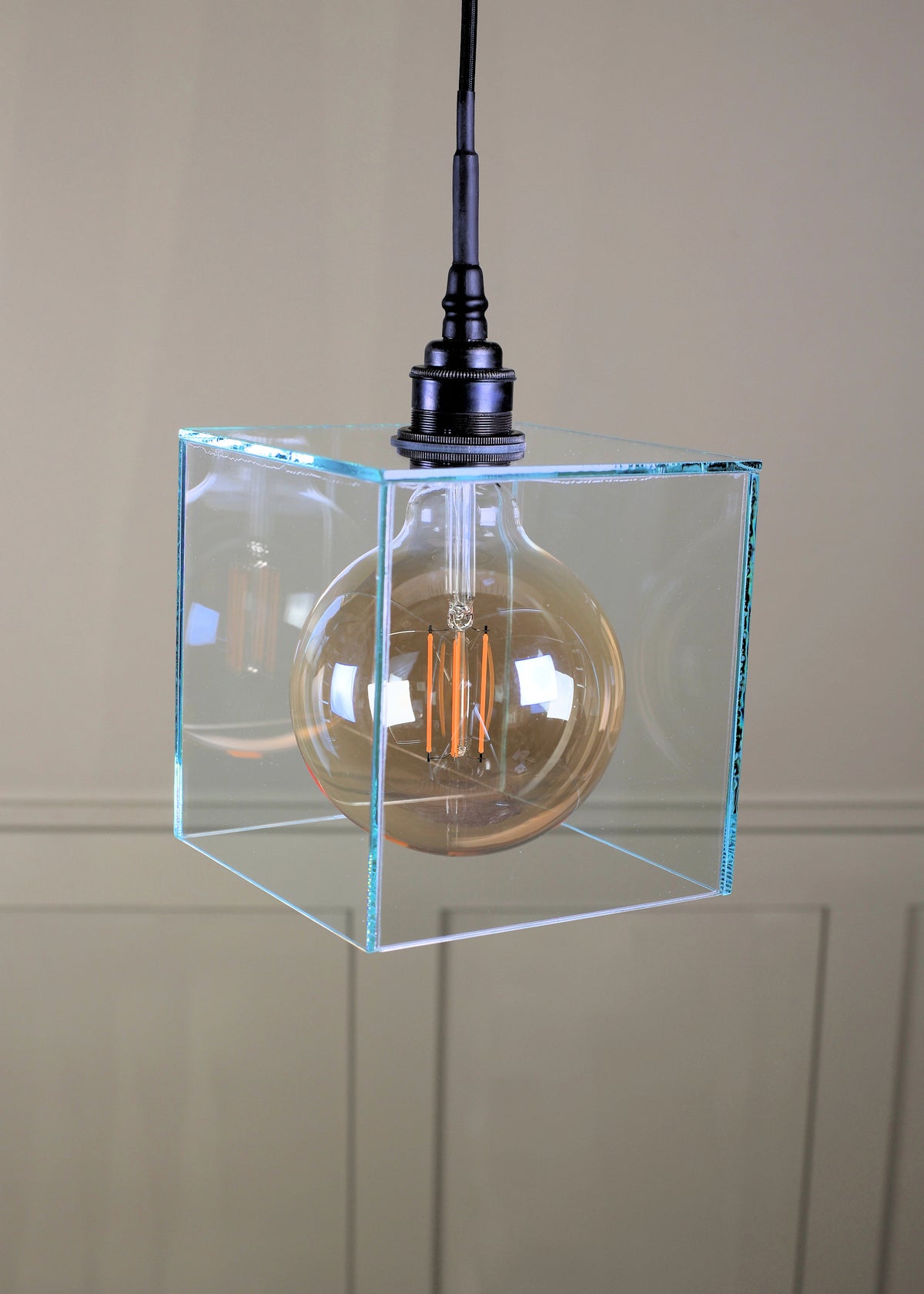 A modern pendant light featuring a clear glass cube shade enclosing a warm-toned Edison bulb, suspended from a black metal ceiling mount with a black cord.