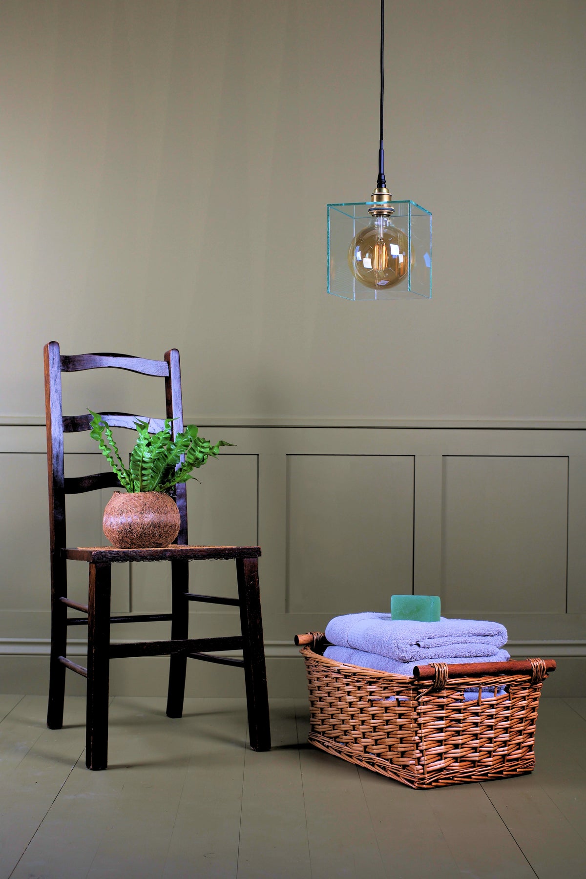 A hanging pendant light with a clear glass cube and visible bulb, with a wooden chair, potted plant, and a wicker basket holding folded blue towels.