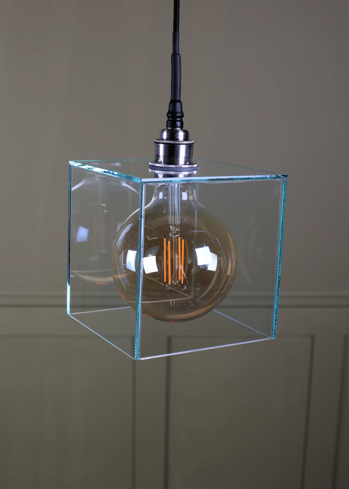 A modern pendant light featuring a clear glass cube frame enclosing a large amber glass globe bulb with a visible filament, suspended by a black cord with a silver metal cap against a neutral paneled wall.