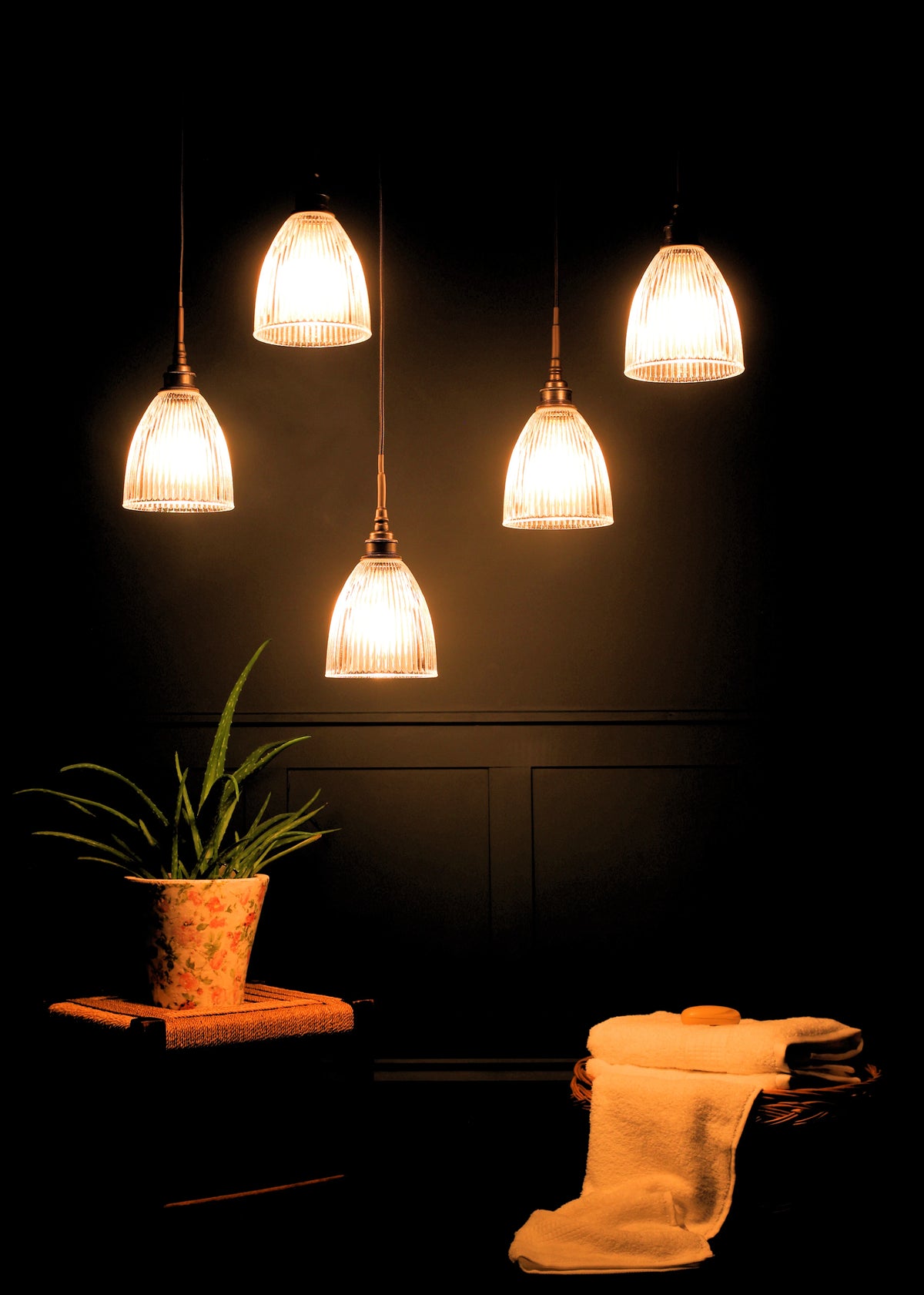 Five pendant light fixtures with ribbed glass shades and dark metal cords hang against a dark teal paneled wall above a small stool with a potted plant and a basket with folded white towels in dark room