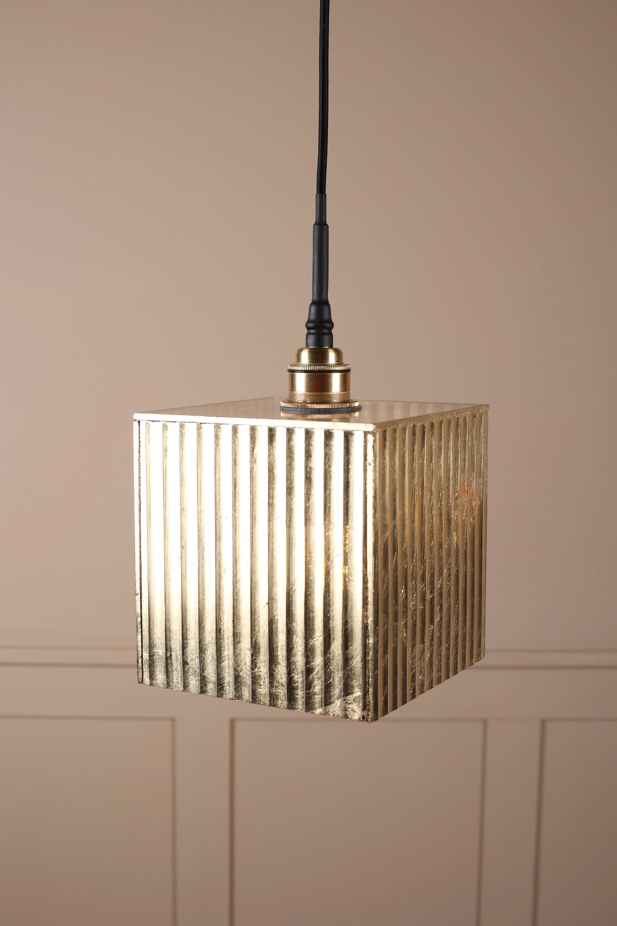 A square pendant light with a ribbed metallic gold shade, suspended by a black cord against a beige paneled wall.