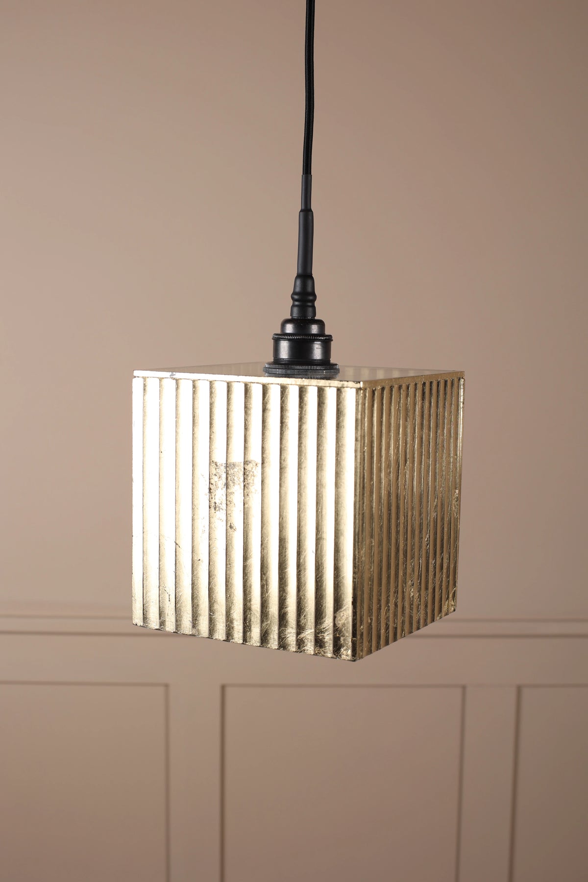 A square pendant light with a textured, vertically ribbed metallic gold shade hanging from a black cord against a beige paneled wall.