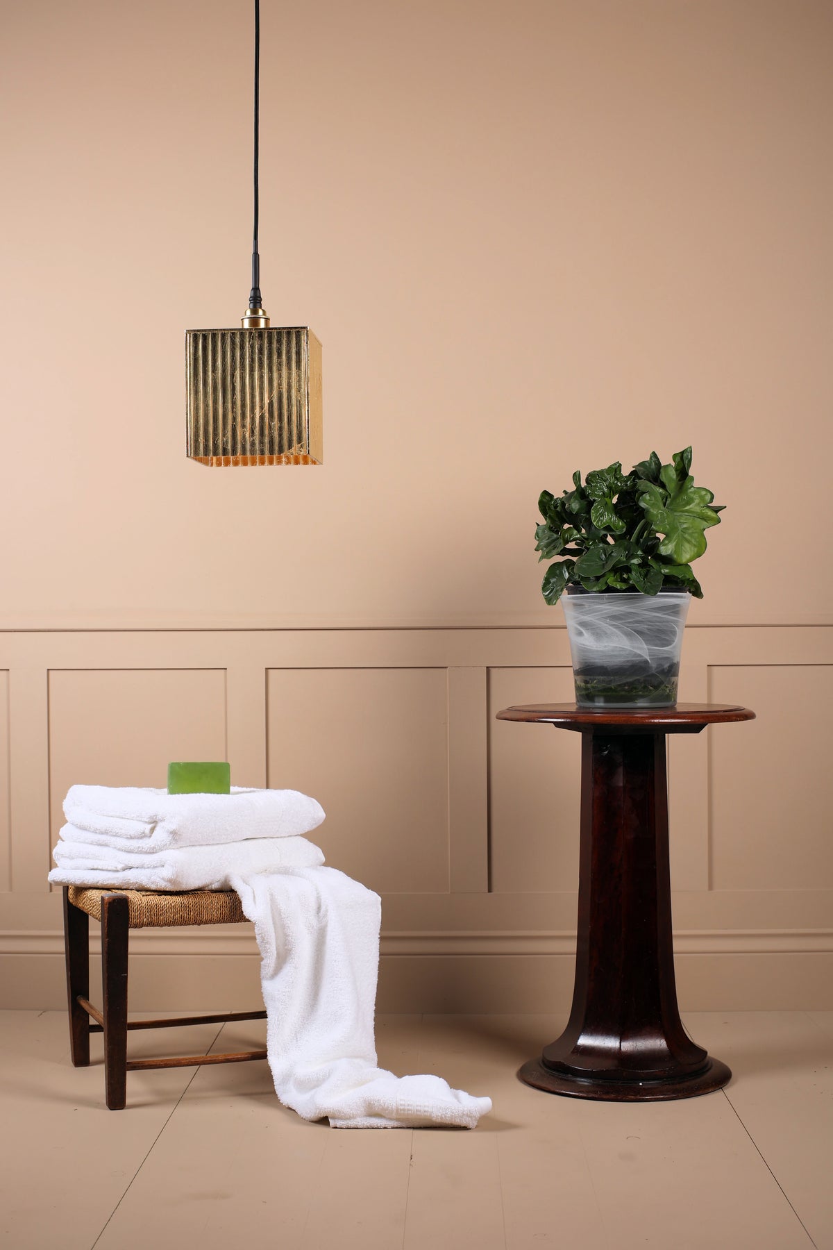 Gold square bathroom pendant light hanging over white towels, wooden table with plant, beige wall